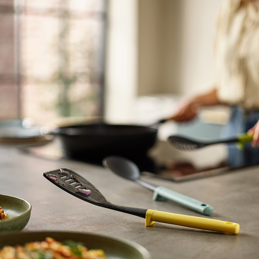 Una escena de cocina con utensilios de cocina y una persona. Una espátula, una cuchara y una cuchara ranurada están en la encimera. Una sartén está en la estufa.