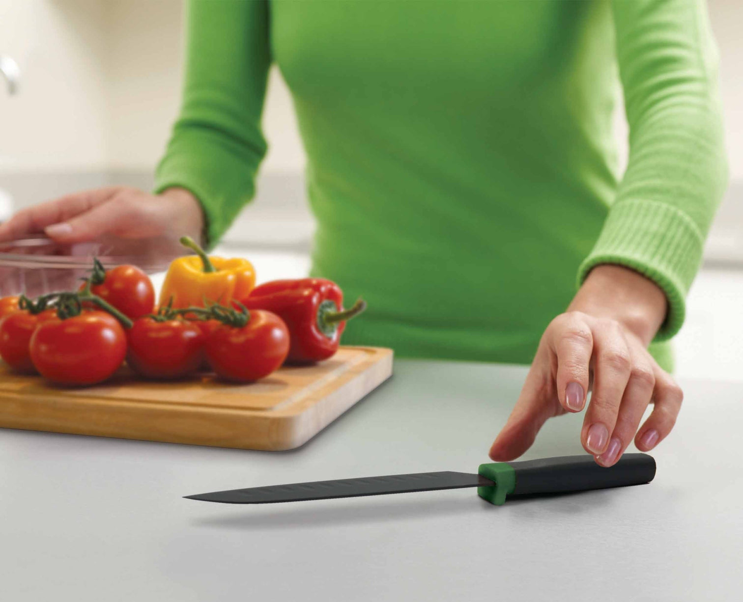 Una persona de verde alcanza un cuchillo negro. Tomates y pimientos en una tabla de cortar. El fondo es una cocina.