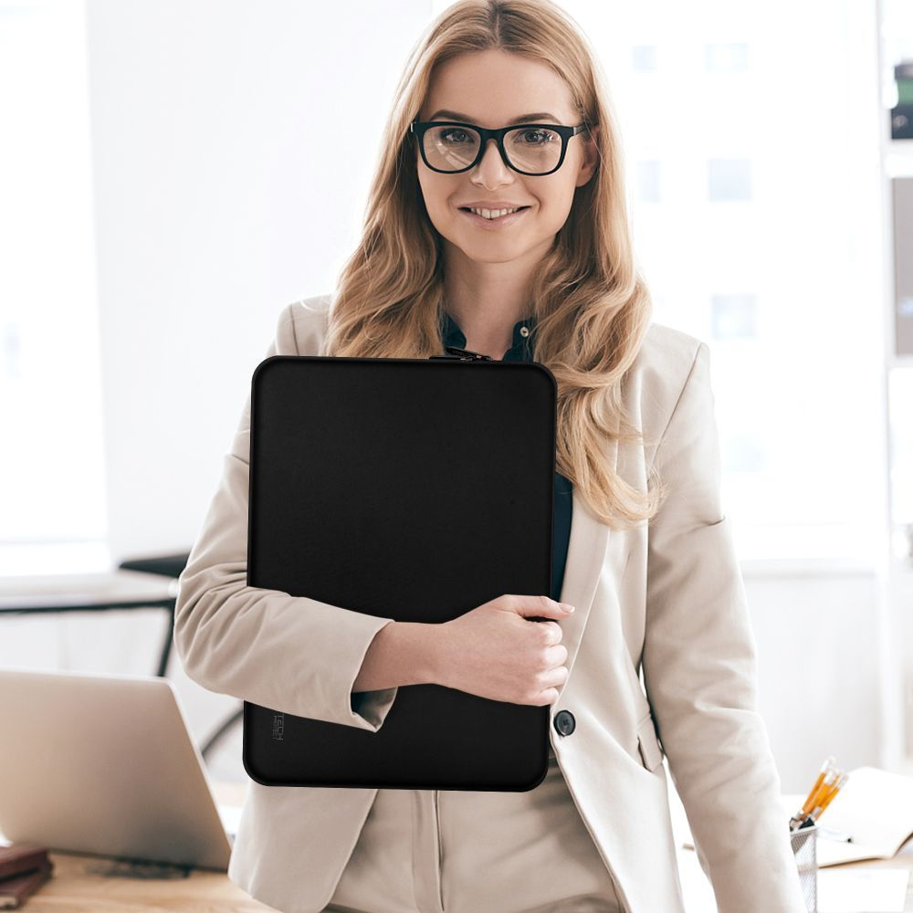 Een vrouw met bril en blazer houdt een zwarte laptoptas vast, lachend. Een bureau met een laptop en kantoorbenodigdheden op de achtergrond.