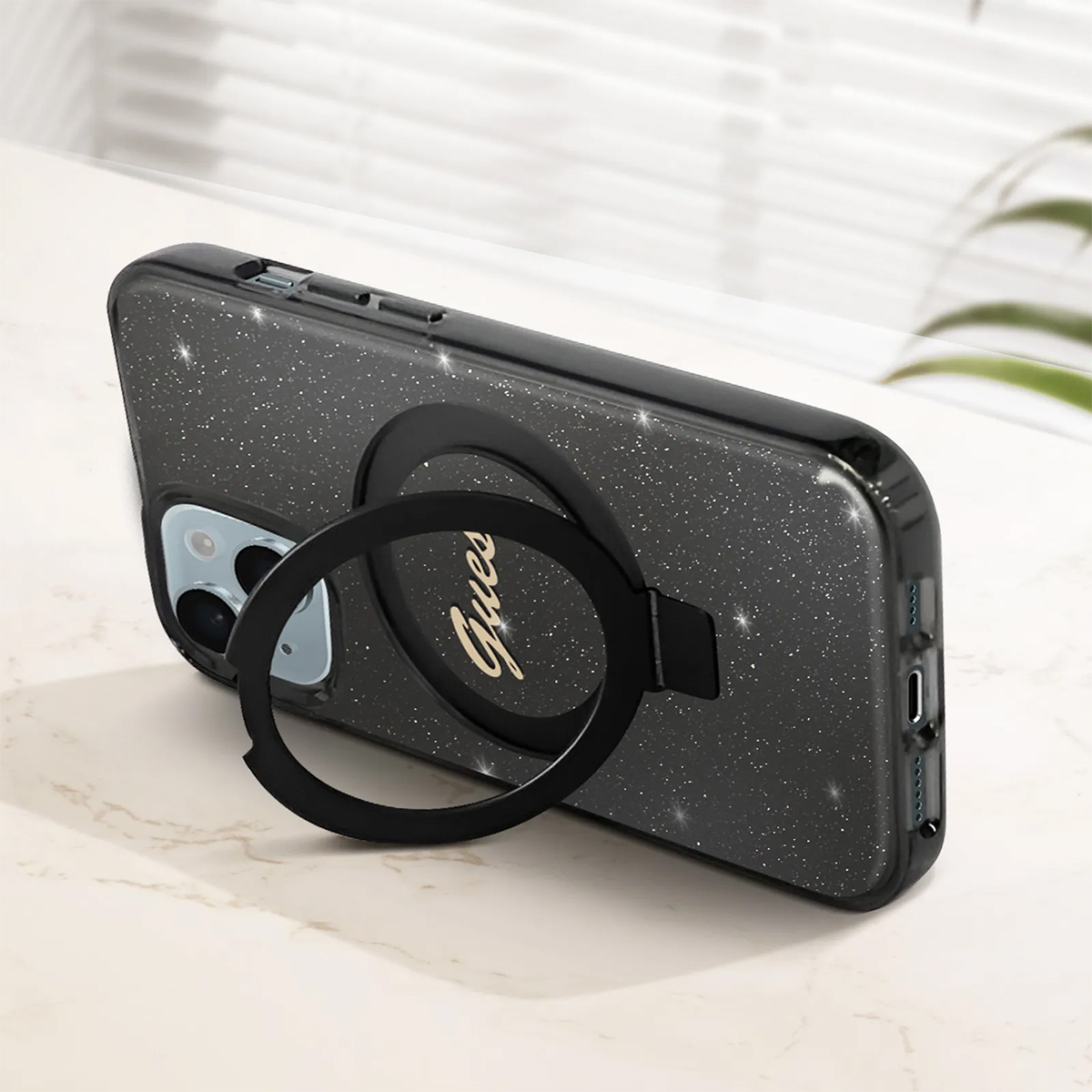 Una custodia per telefono nera glitterata con un anello nero, con il logo 'Guess', poggia su una superficie di marmo. 