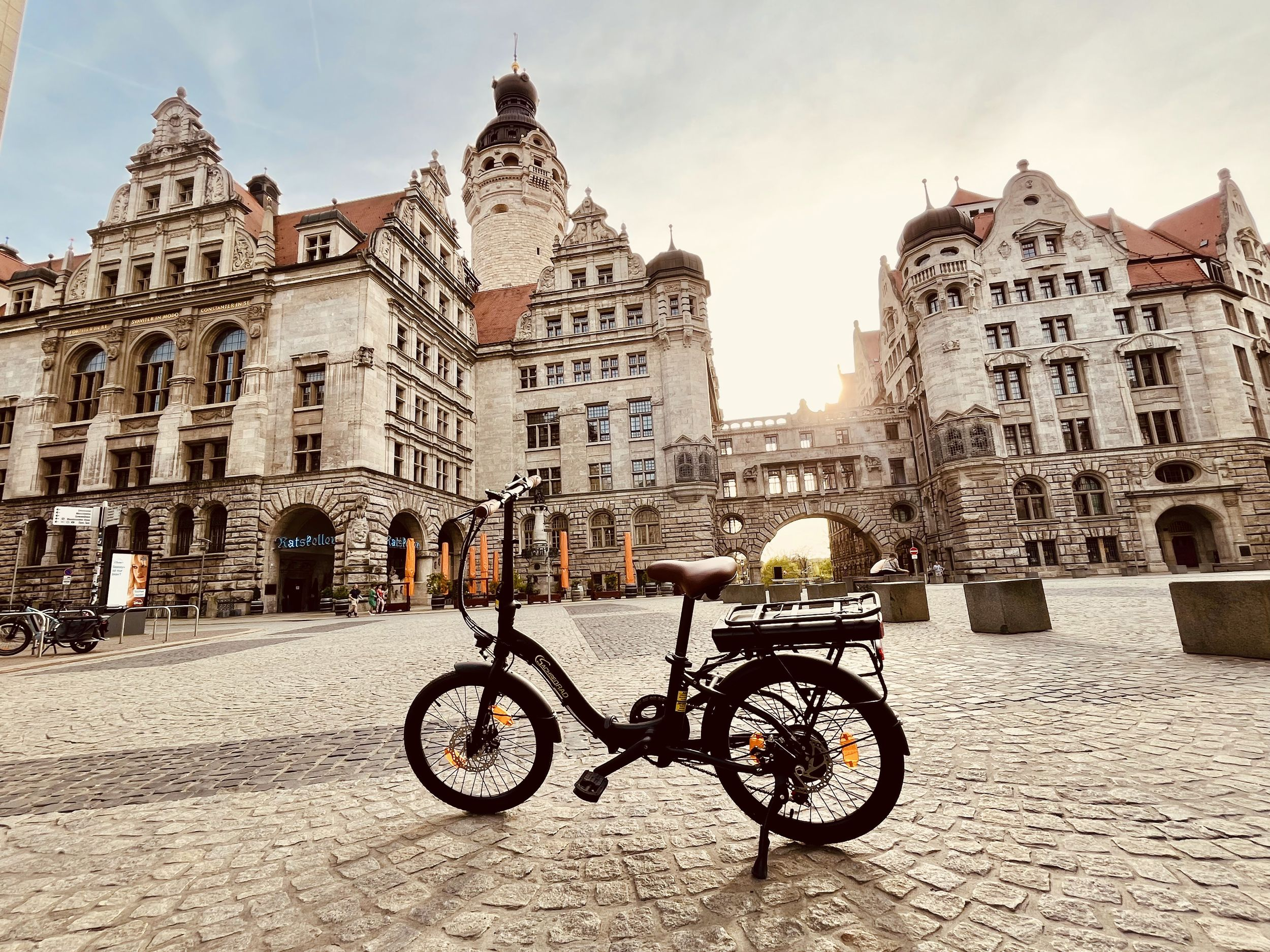 Ein schwarzes Elektrofahrrad steht auf Kopfsteinpflaster vor einem großen, reich verzierten Gebäude mit Turm. Die Sonne scheint.