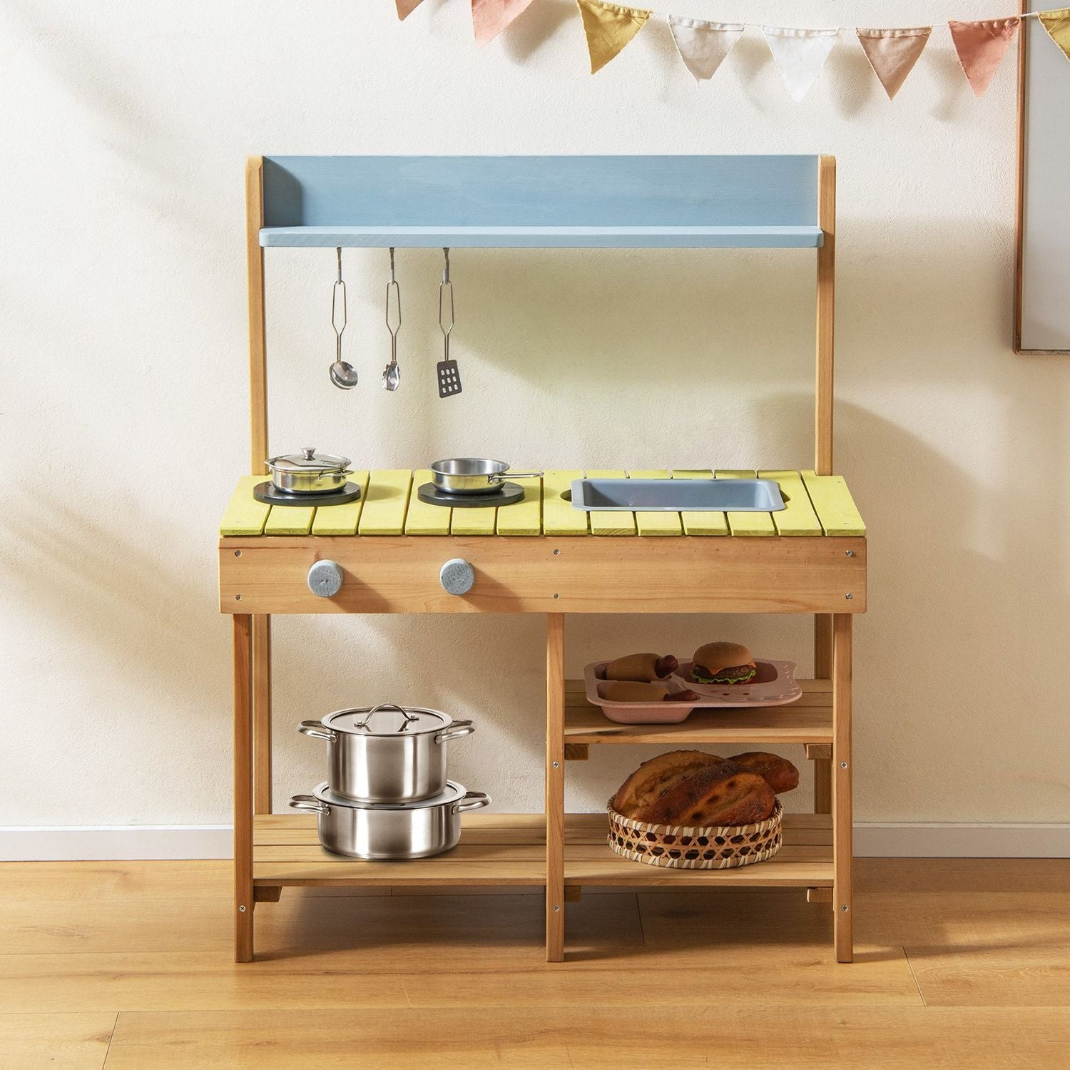 Cocina de juguete de madera con parte superior azul, mostrador amarillo, ollas, sartenes, utensilios y comida de juguete en estantes. Guirnalda arriba.