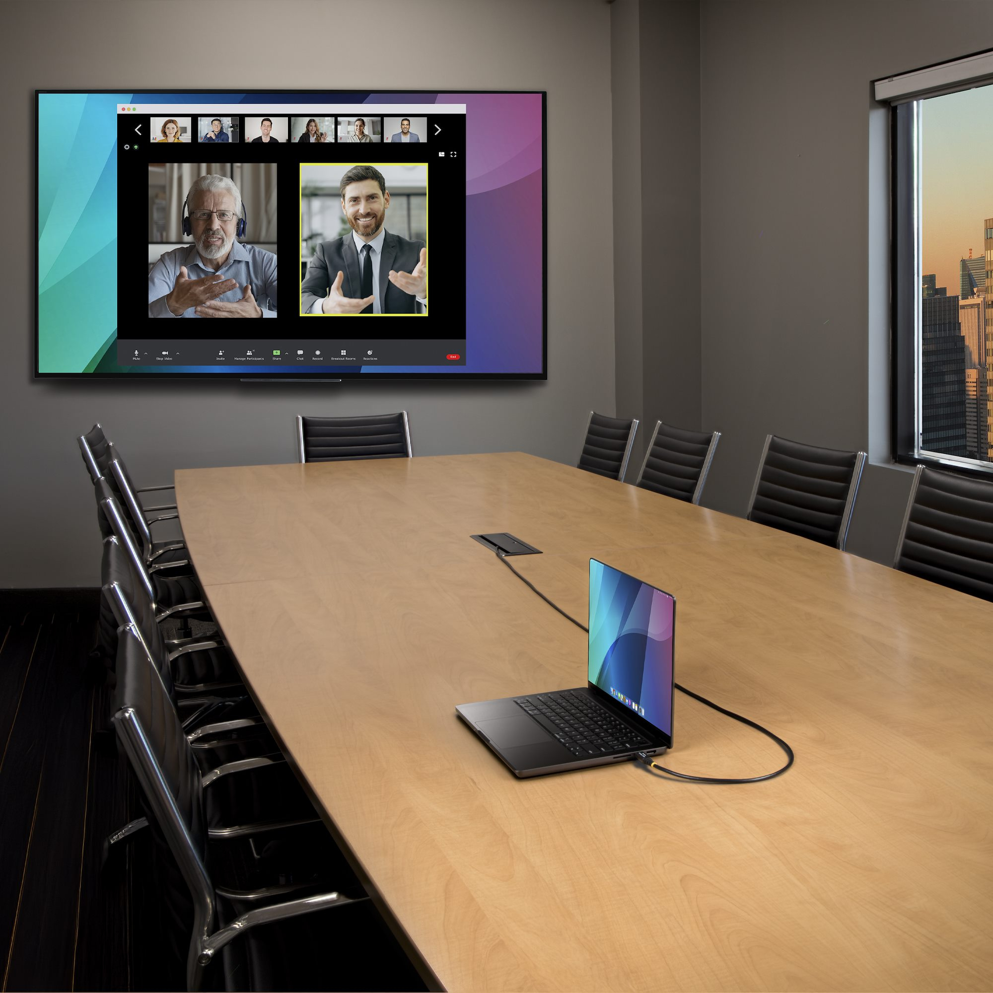 Konferenzraum mit Großbildschirm, der einen Videoanruf zeigt. Ein Laptop ist mit dem Bildschirm verbunden, der Tisch hat Stühle.