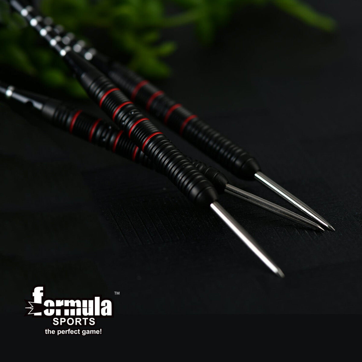 Nahaufnahme von drei schwarzen und roten Dartpfeilen mit silbernen Spitzen. Formula Sports Logo unten. Dunkler Hintergrund mit grünen Blättern.