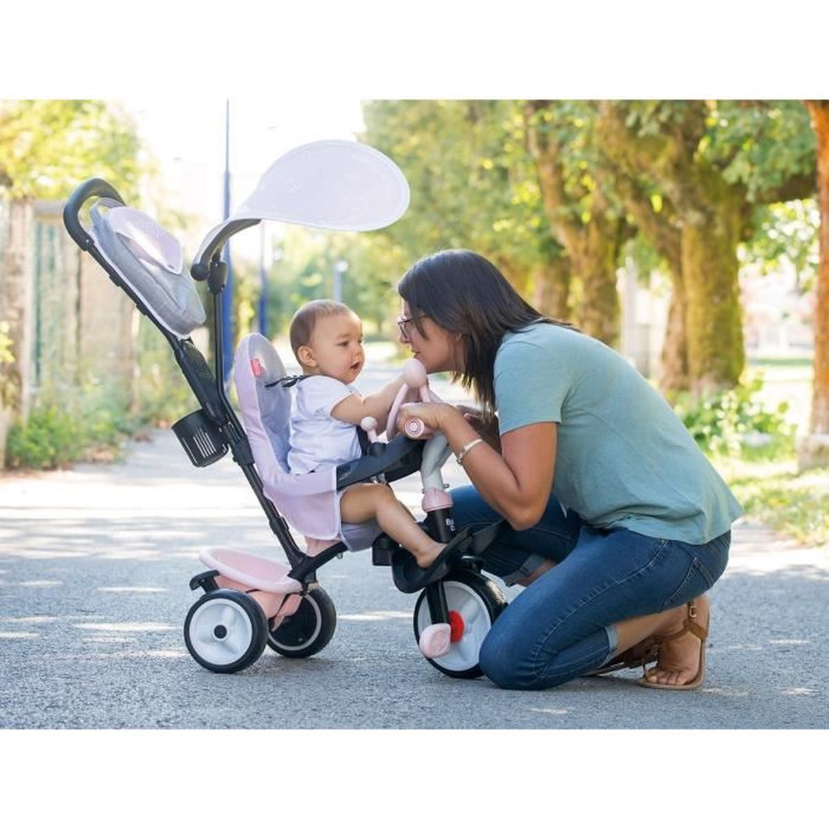 Una donna e un bambino interagiscono con un triciclo rosa. La donna si inginocchia, guardando il bambino. Gli alberi costeggiano lo sfondo.