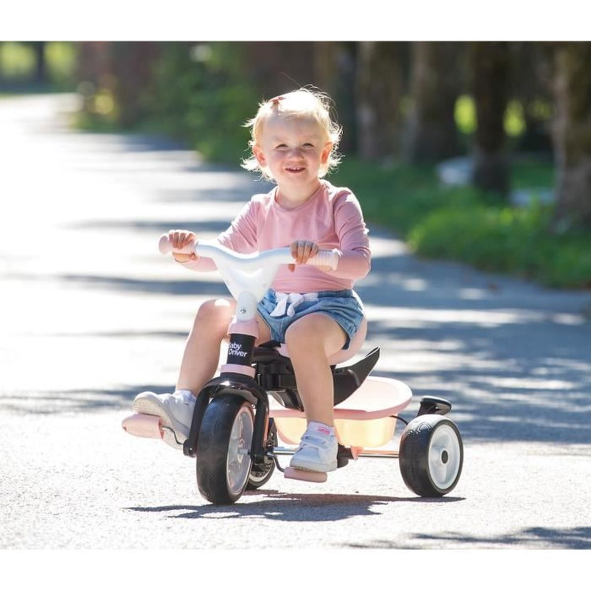 Un bambino sorridente guida un triciclo rosa su un sentiero pavimentato, indossa una maglietta rosa e pantaloncini di jeans, alberi sullo sfondo.