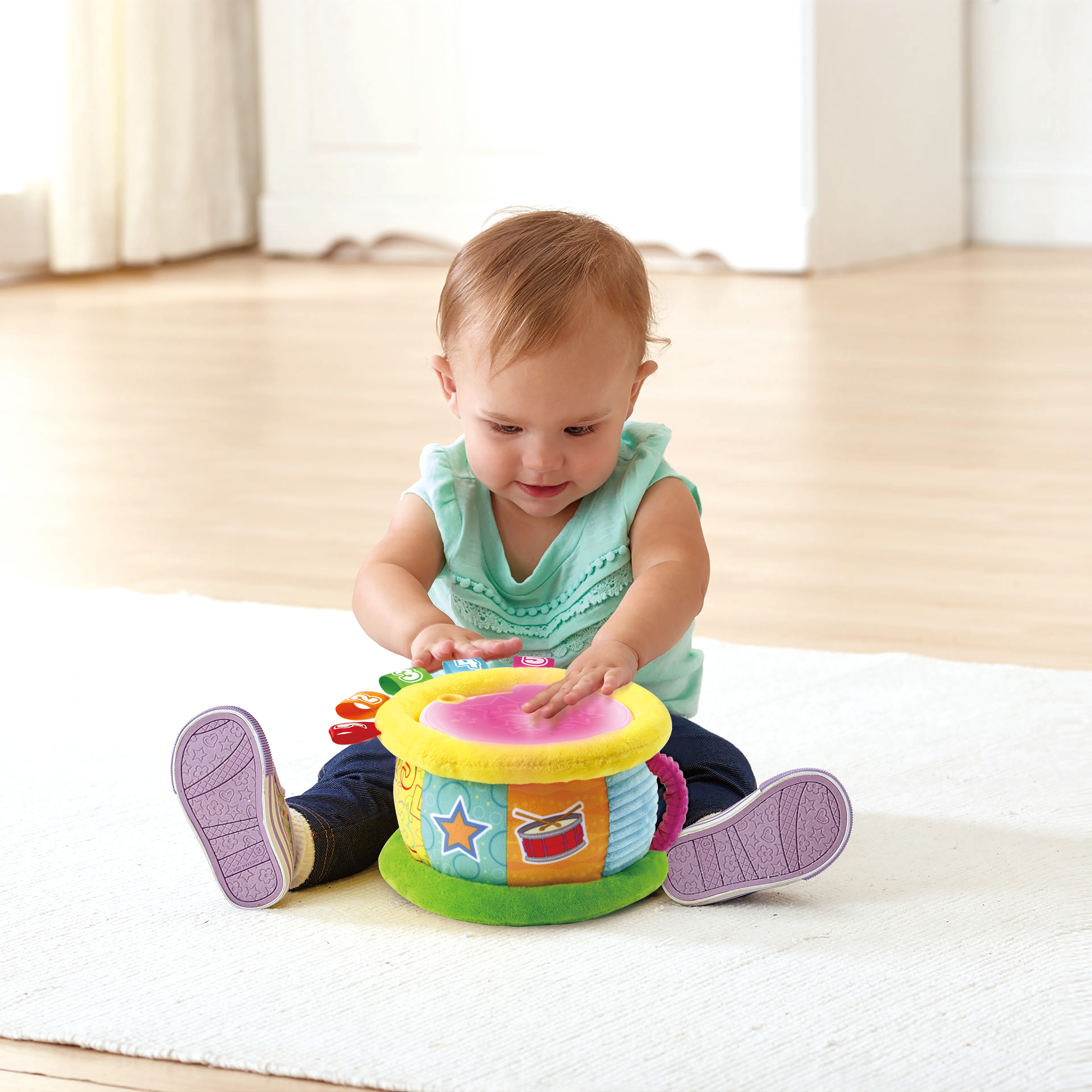 Ein Baby spielt auf einer bunten Spielzeugtrommel. Das Baby trägt ein blaues Hemd und Jeans. Die Trommel steht auf einem weißen Teppich.
