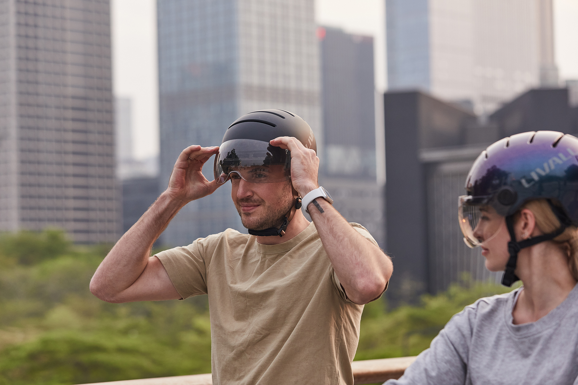 Un uomo regola la visiera del casco. Una donna con il casco accanto a lui. Edifici della città sullo sfondo. Cespugli verdi.