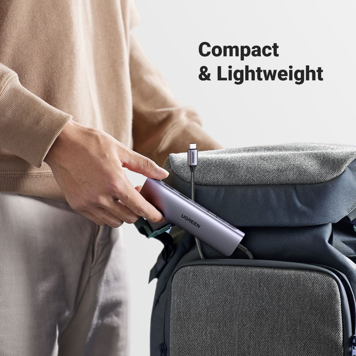 Eine Person legt einen UGREEN USB-C-Hub in einen Rucksack. Der Hub ist silberfarben und mit einem Kabel verbunden. Text: 'Compact & Lightweight'.