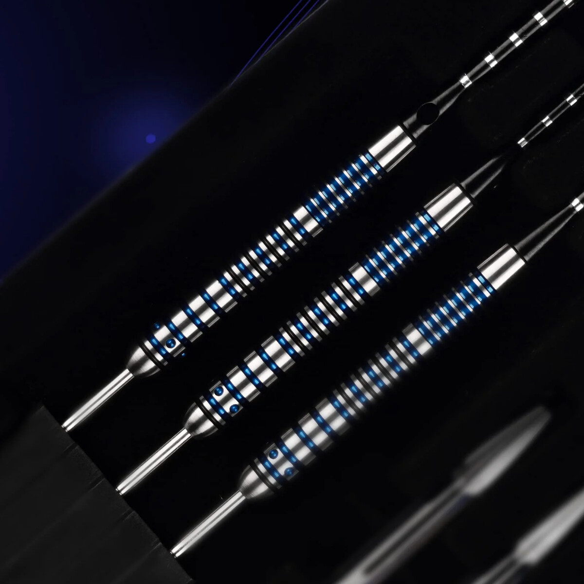 Close-up of three darts with silver bodies and blue accents, arranged in a dark case against a blurred black background.