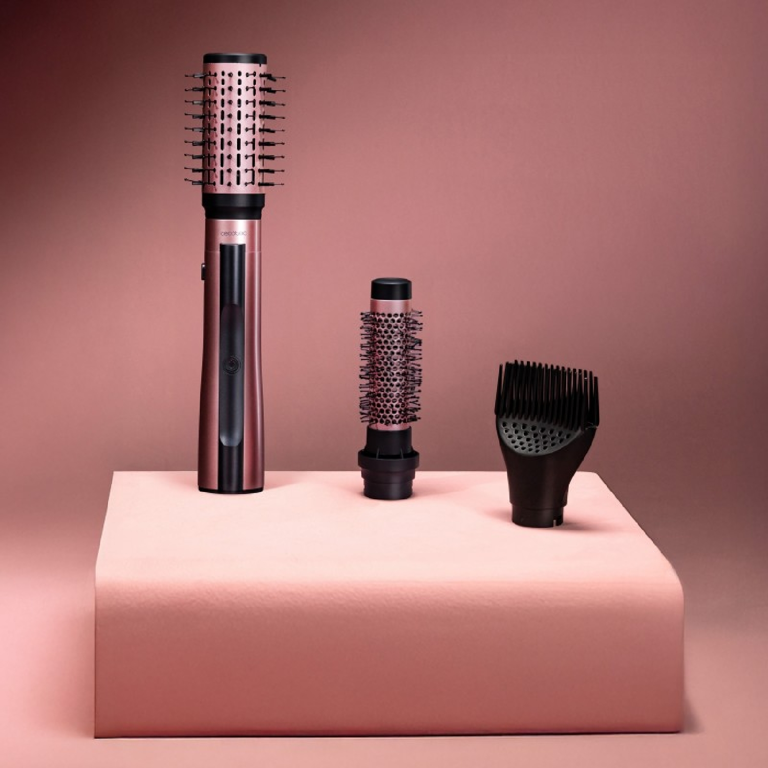 Drei roségoldene Haarstyling-Tools auf einer rosa Plattform. Enthält eine Bürste, eine kleinere Bürste und eine Düse, alles vor einem rosa Hintergrund.