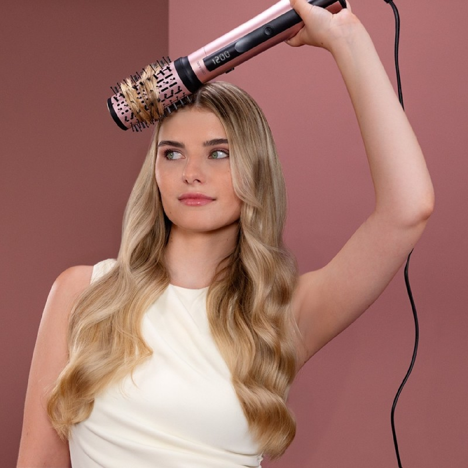 Una donna usa una spazzola per capelli. Ha lunghi capelli biondi e indossa un vestito bianco. Lo sfondo è rosa.