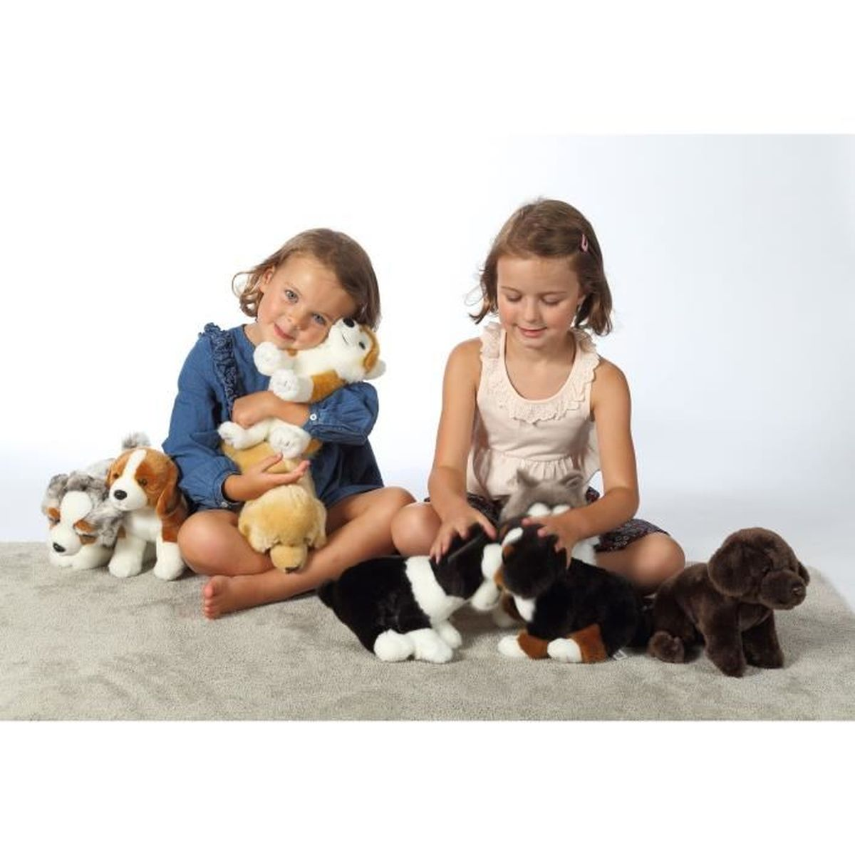 Zwei Mädchen sitzen mit Stoffhundespielzeugen auf einem Teppich. Sie sind von Plüschhunden verschiedener Farben und Rassen umgeben.
