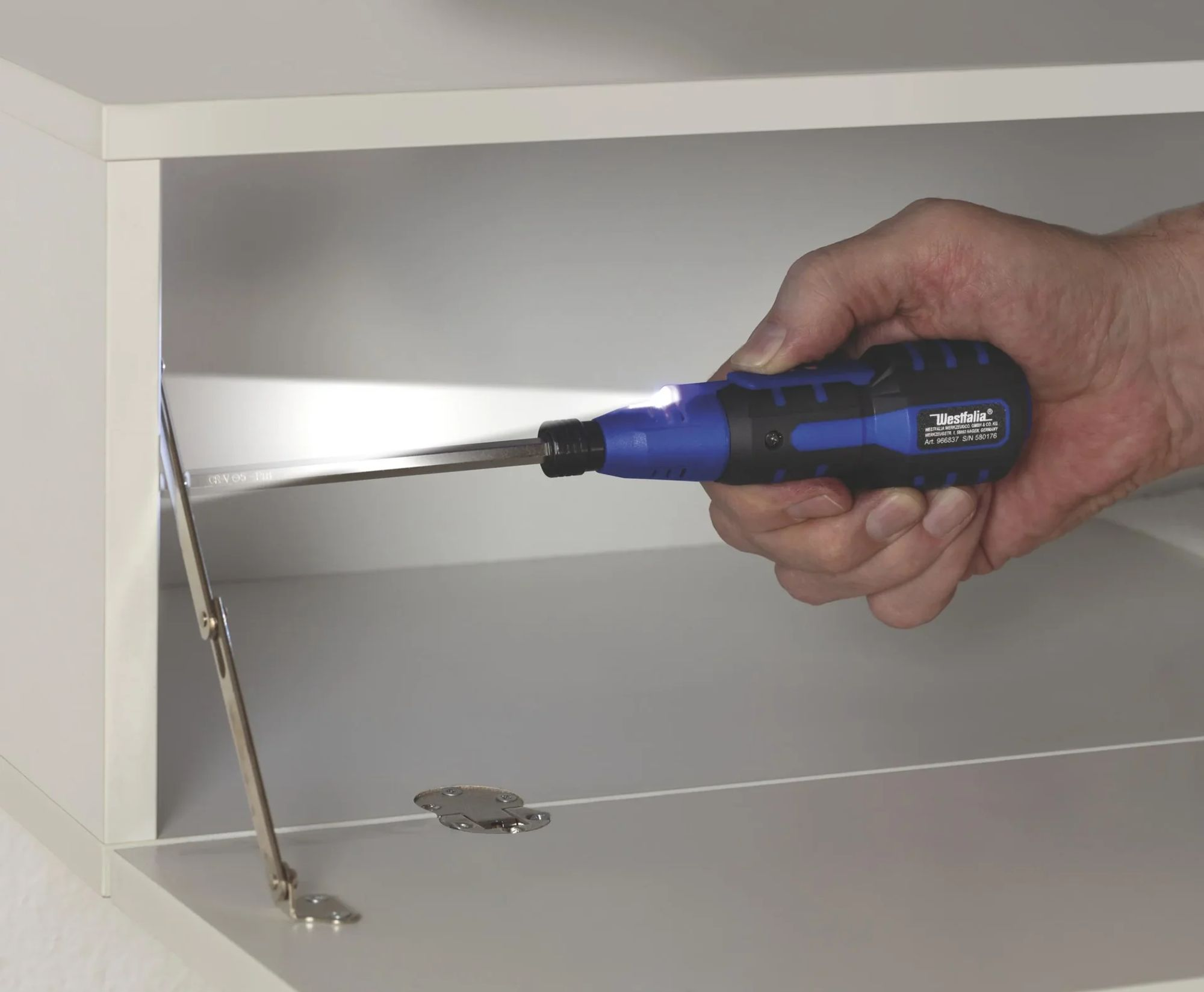 Eine Hand hält einen blau-schwarzen Schraubendreher mit Licht und zeigt ihn in einen offenen weißen Schrank.