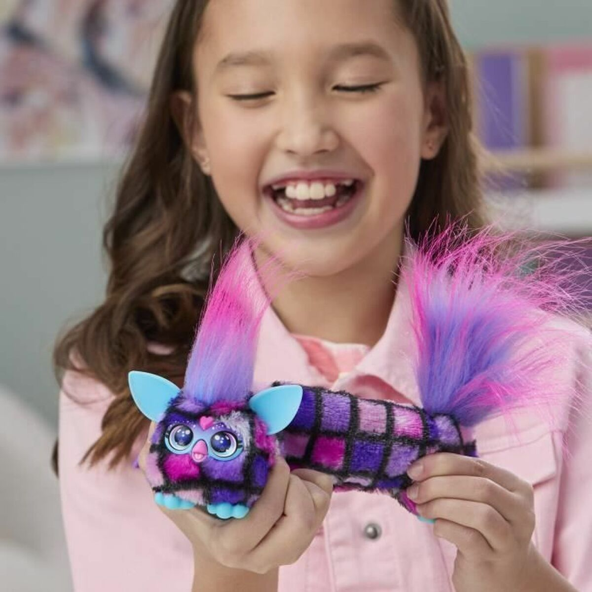 Ein lächelndes Mädchen hält ein farbenfrohes Furby-Spielzeug. Das Spielzeug hat rosa, lila und blaue Haare. Das Mädchen trägt ein rosa Hemd.