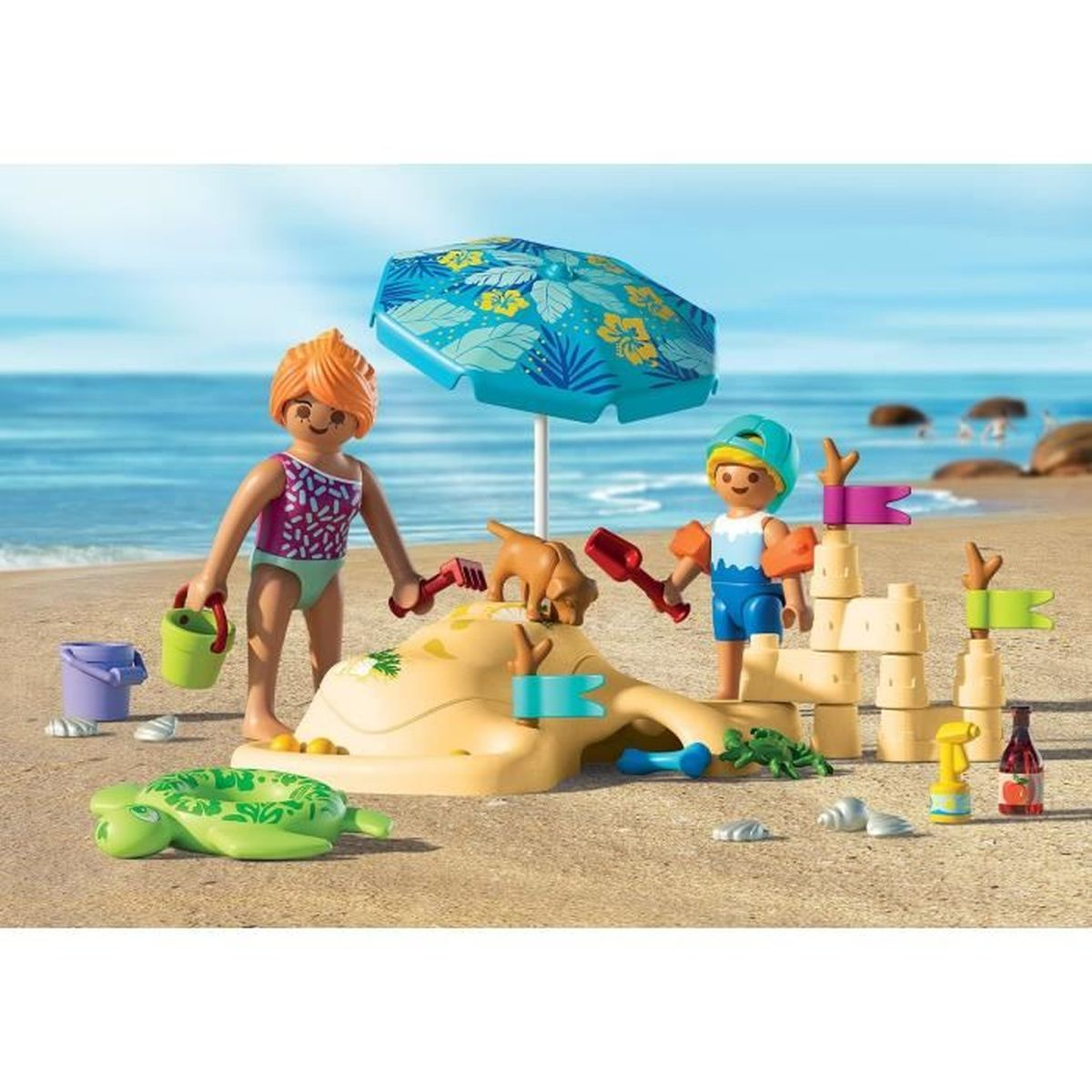 Scena sulla spiaggia con personaggi Playmobil, castello di sabbia, cane, ombrellone e accessori. Due figure giocano con sabbia e giocattoli.