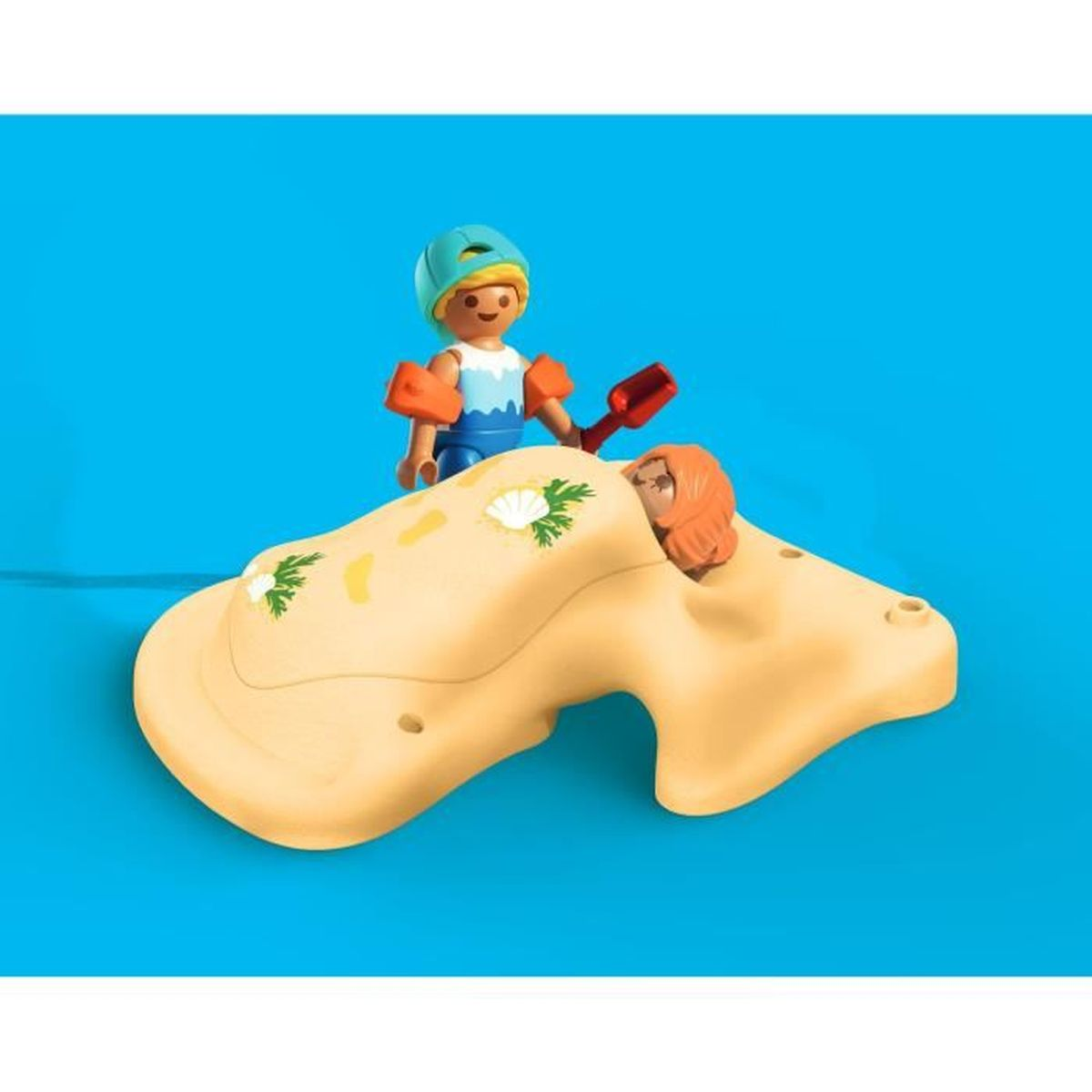 Una figura giocattolo in tenuta da bagno si trova sopra un'altra coperta da una coperta color sabbia, con una pala rossa. Sfondo turchese.