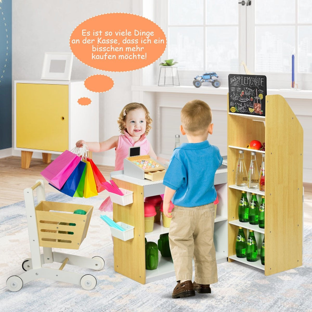 Dos niños juegan a la tienda. Uno sostiene bolsas de compras. La tienda tiene caja registradora, estantes con botellas y juguetes. Un niño compra.