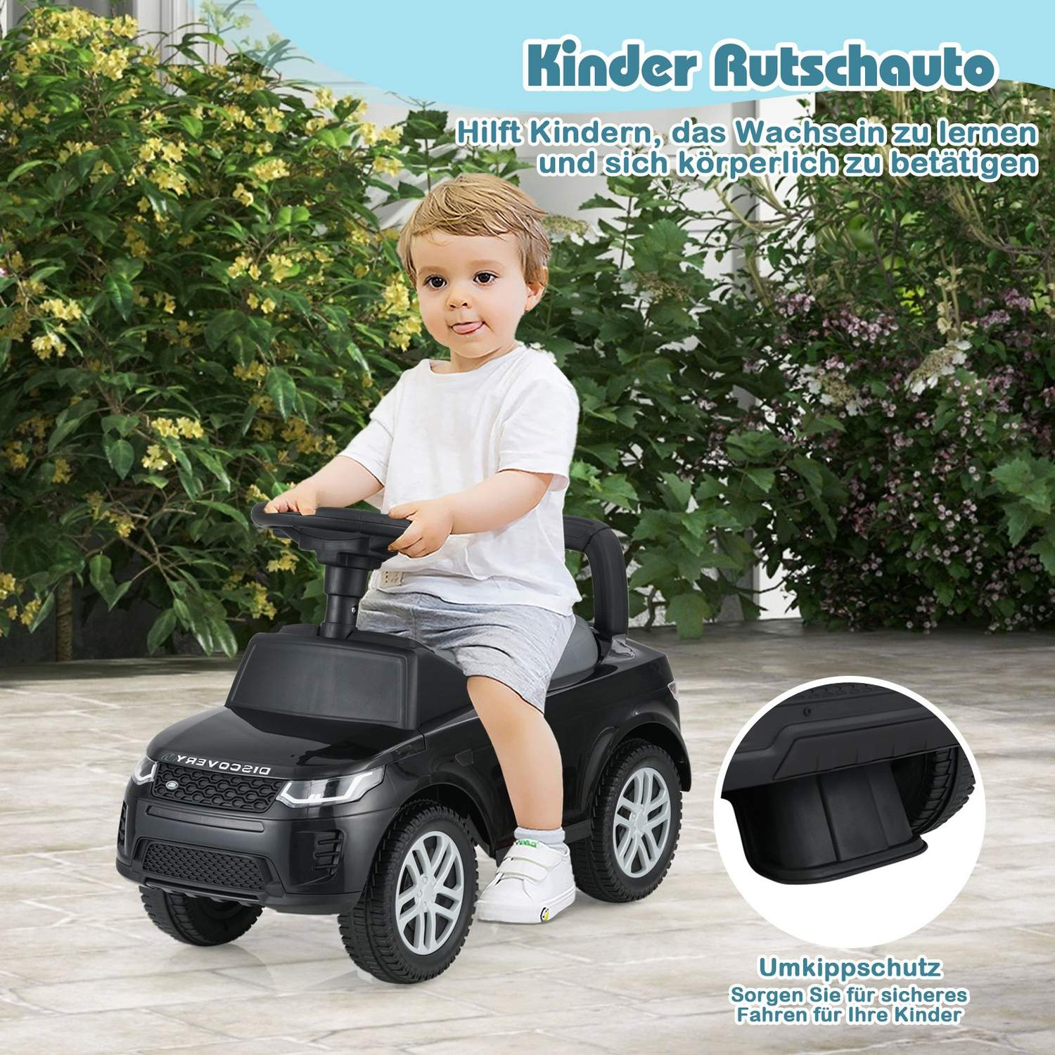 Un niño pequeño está sentado en un coche negro. El coche está en un patio con arbustos verdes en el fondo.