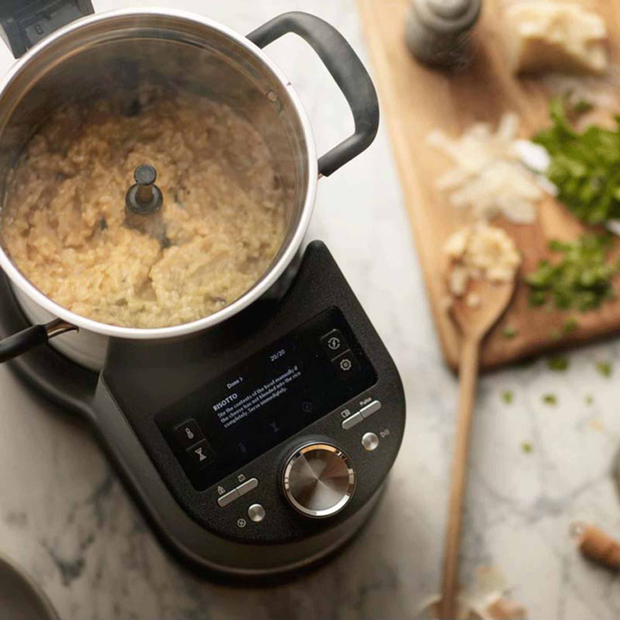 Risotto kocht in einem Topf auf einer schwarzen Kochmaschine. Zutaten wie Käse und Petersilie liegen auf einem Holzbrett.