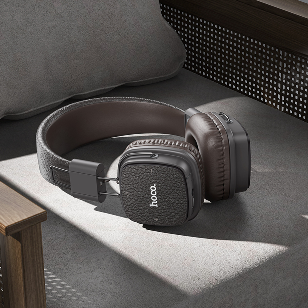 Auriculares Hoco marrones y negros sobre una superficie gris, junto a una almohada y muebles de madera. Los auriculares son el foco.