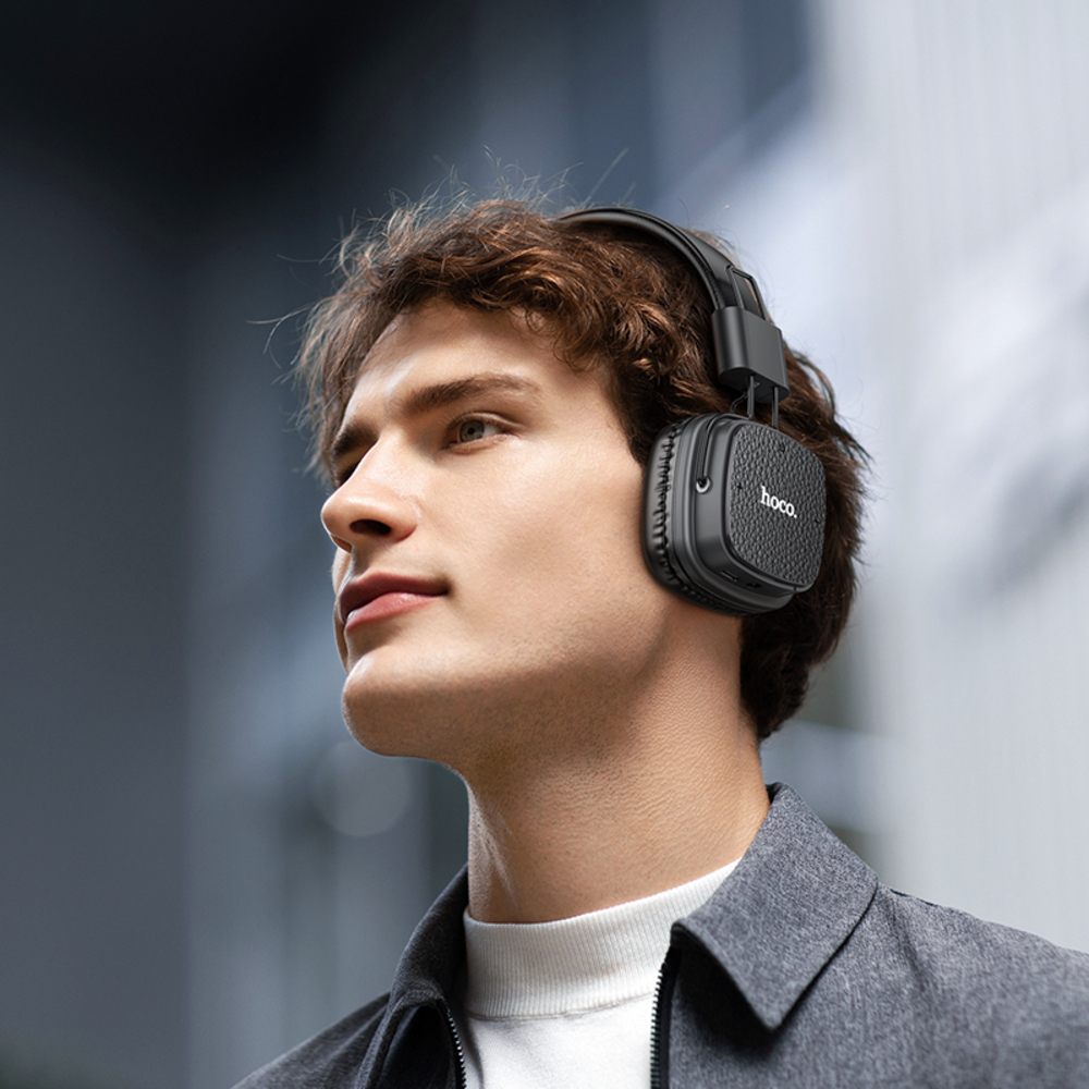 Un hombre con auriculares negros mira hacia arriba. Los auriculares tienen el logo 'hoco.' en ellos. Lleva una chaqueta gris y una camisa blanca.
