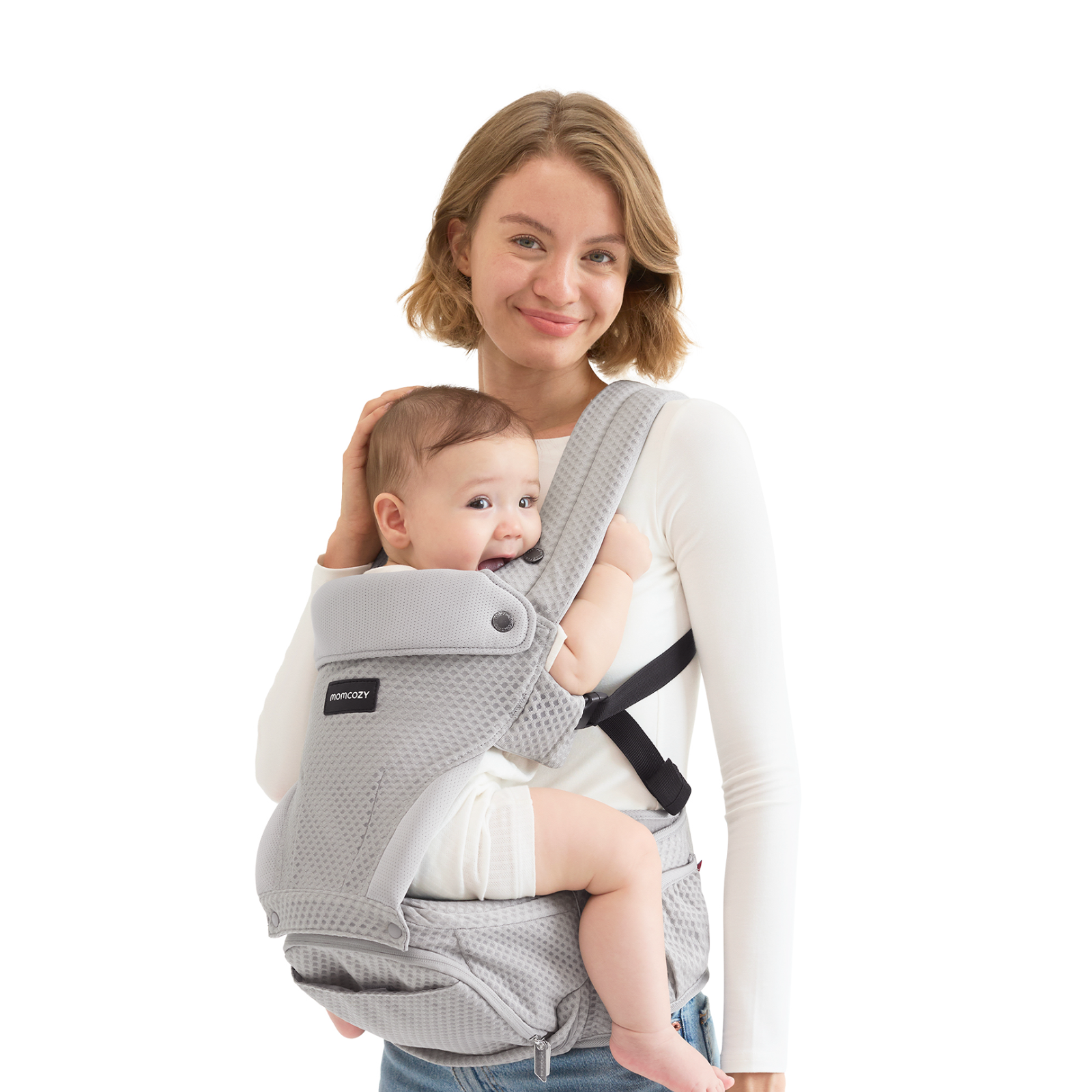 A woman holds a baby in a gray carrier. Both are in front of a white background. The woman smiles.