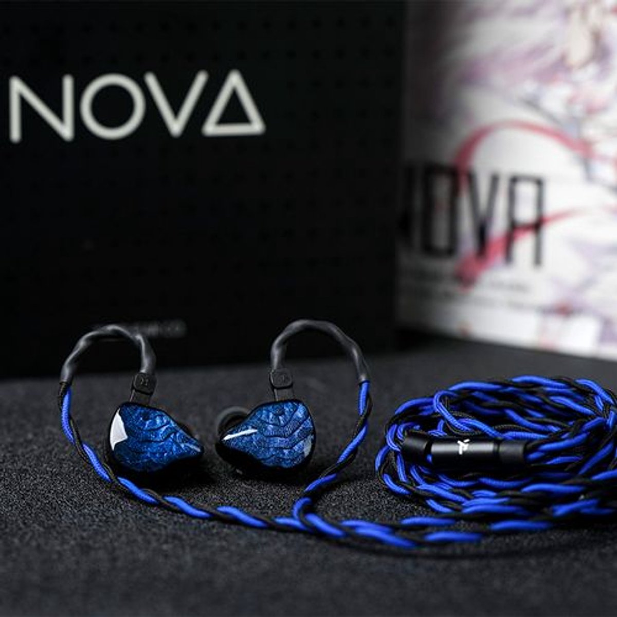 Oordopjes met blauwe en zwarte kabel, naast een zwarte doos met de tekst "NOVA". Op de achtergrond staat een witte en roze doos.