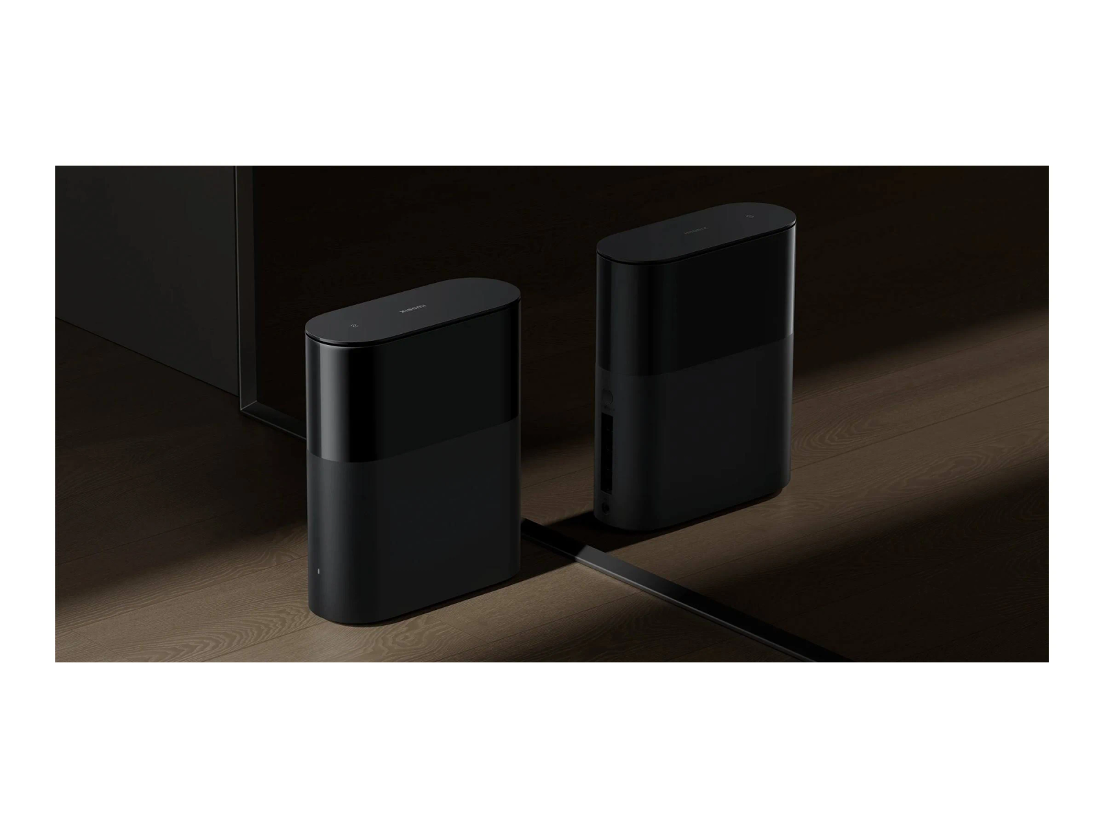 Two black speakers on wooden floor. Each has a dark top and a lighter bottom section. The background is dark.