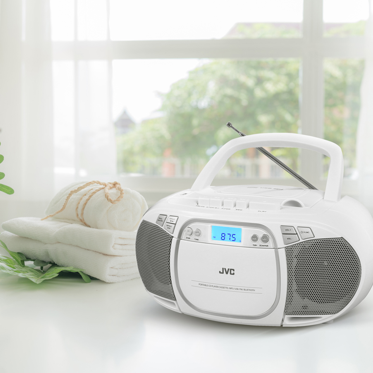 Un boombox JVC bianco con display digitale 87,5 è su un tavolo bianco vicino a asciugamani piegati e una foglia verde.