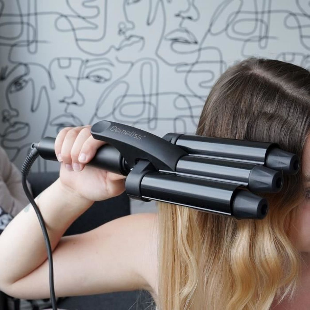 Eine Frau benutzt einen schwarzen Haarwellenstab auf ihrem blonden Haar. Der Hintergrund zeigt eine gemusterte Wand mit Gesichtern.