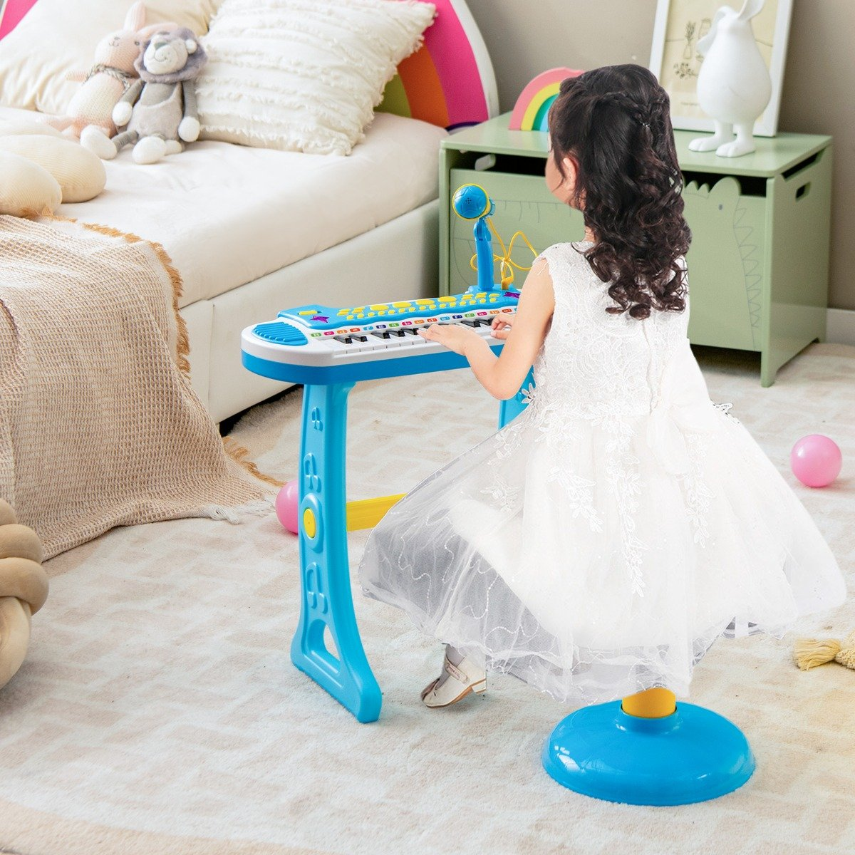 Una niña con un vestido blanco toca un piano de juguete azul. La habitación tiene una cama, juguetes y una decoración de arcoíris.