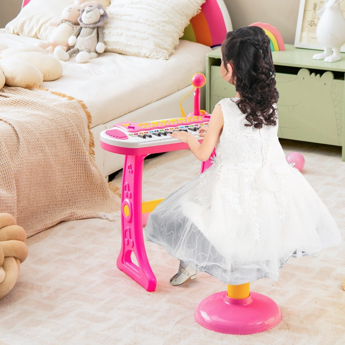 Una niña con vestido blanco toca un piano de juguete rosa. Un micrófono está cerca. Juguetes y una cama en el fondo.