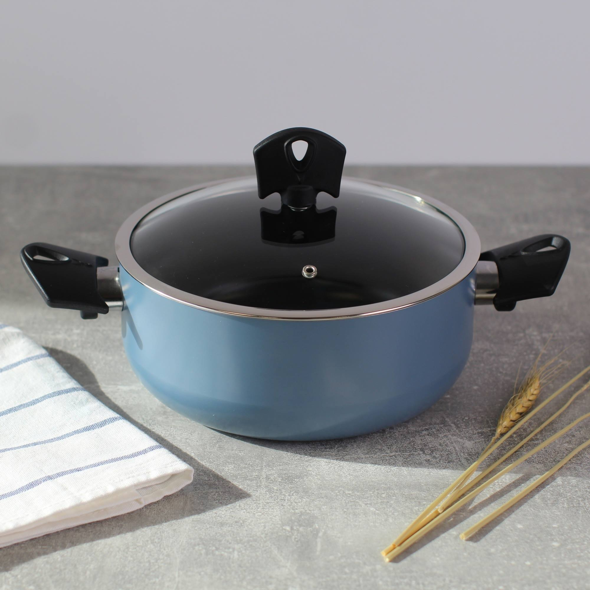 Una olla azul con tapa de cristal y asa negra sobre una superficie gris, junto a un paño blanco y trigo.