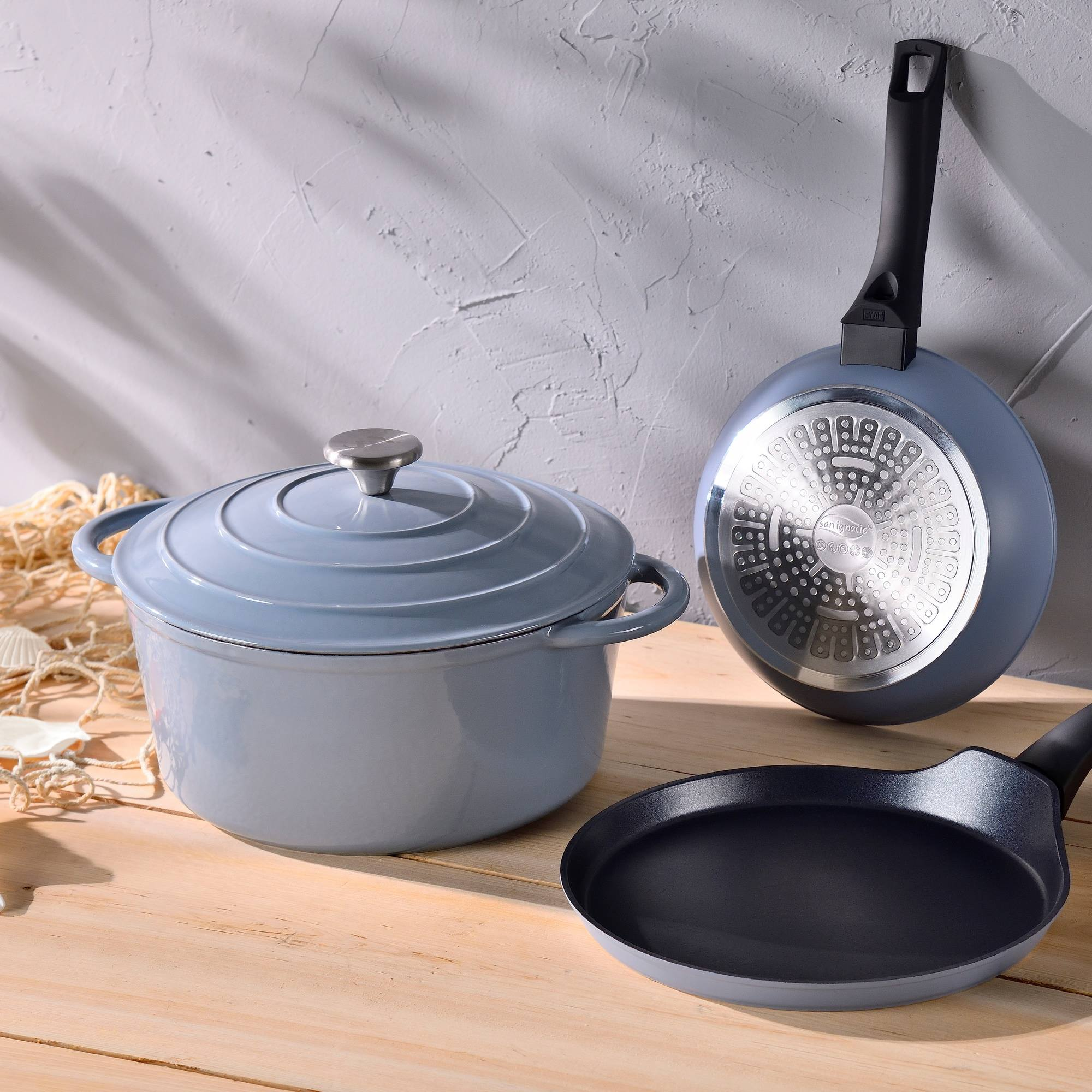 Tres utensilios de cocina: una olla azul con tapa, una sartén y otra sartén, sobre una superficie de madera.