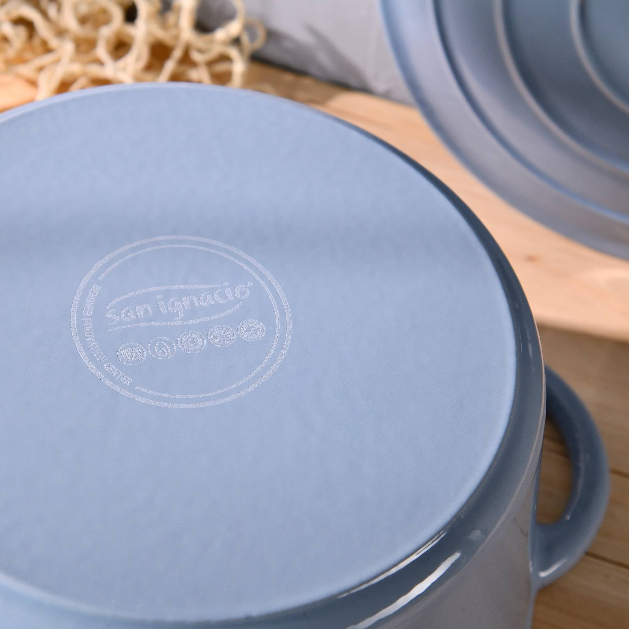 Primer plano de una olla azul claro con el logotipo de San Ignacio. El fondo incluye superficie de madera y platos.