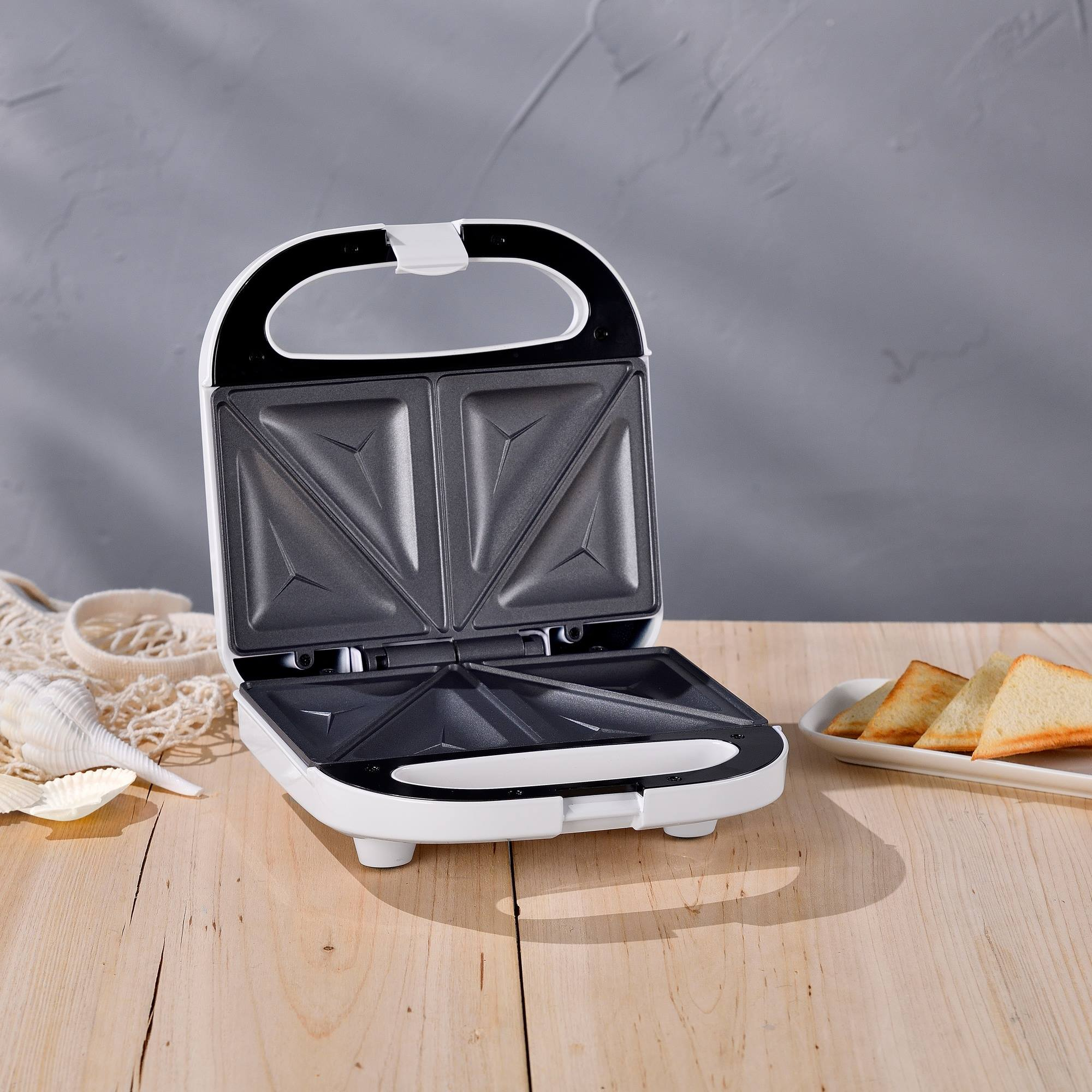 Una sandwichera blanca está abierta sobre una mesa de madera. Rebanadas de tostadas en un plato blanco. Una bolsa de red con conchas.