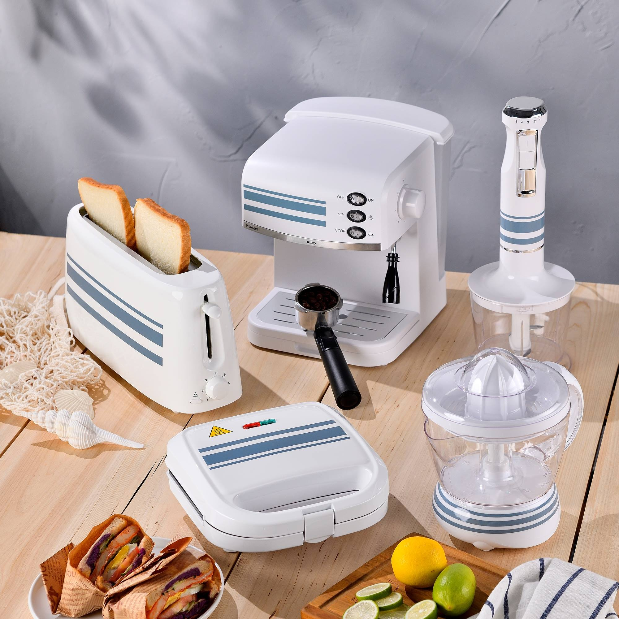 Electrodomésticos de cocina blancos: cafetera, tostadora con pan, exprimidor, sandwichera y batidora sobre una mesa de madera.