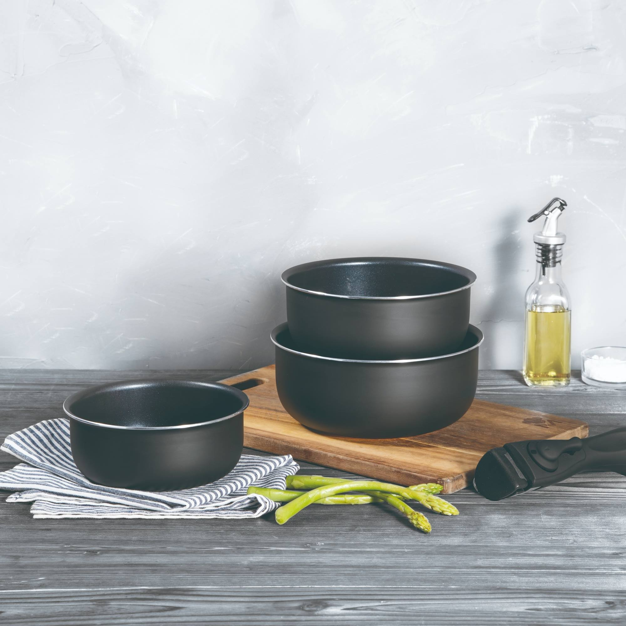 Una escena de cocina con ollas negras, espárragos, aceite y una tabla de cortar de madera sobre una superficie de madera gris.