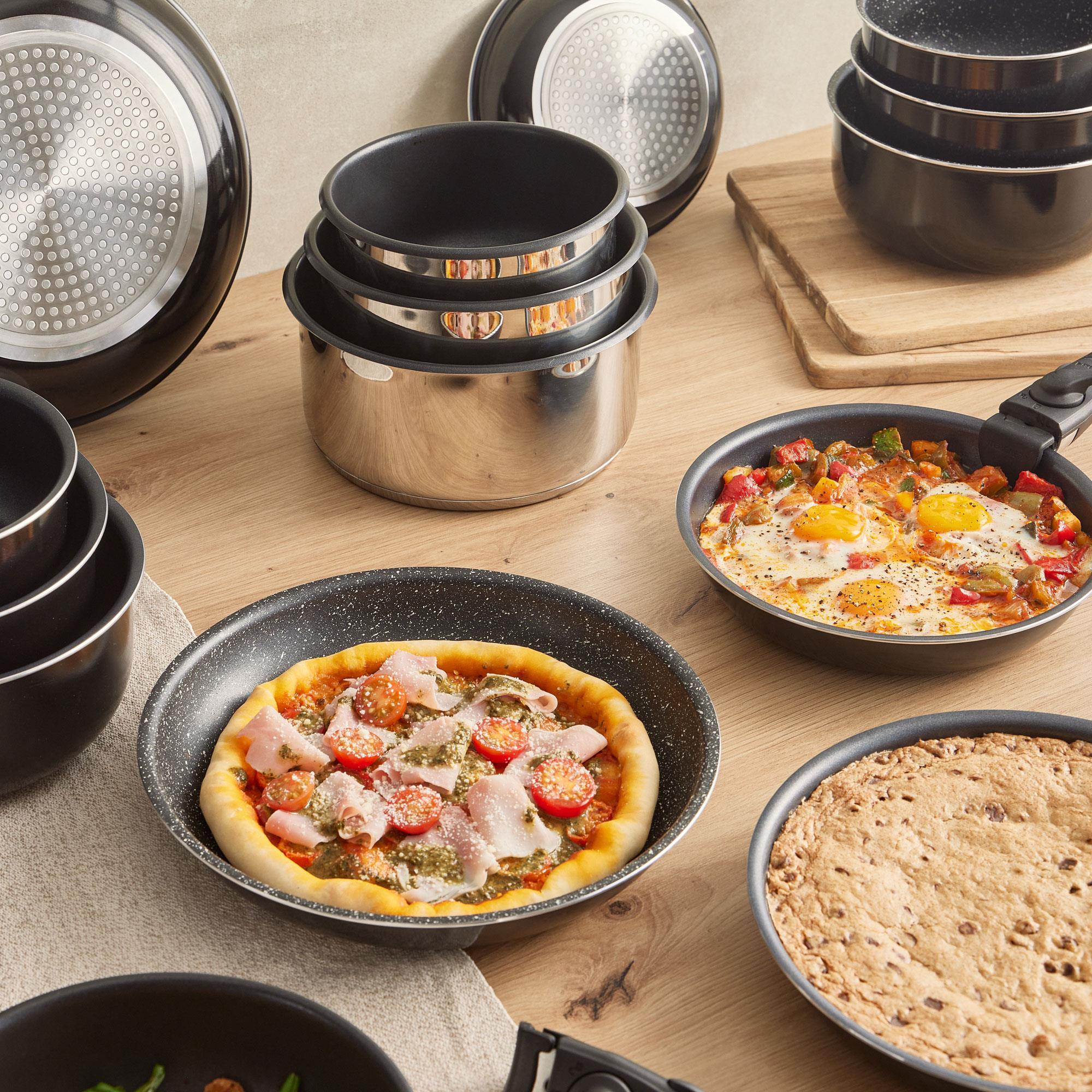 Una mesa con sartenes, pizza y platos cocinados. Hay ollas y sartenes. Una pizza con tomates.