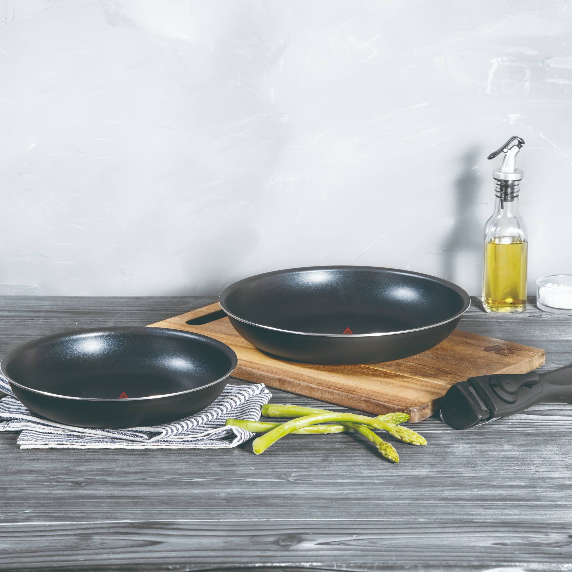 Dos sartenes negros, espárragos y botella de aceite sobre madera. Fondo gris. Ambiente de cocina.
