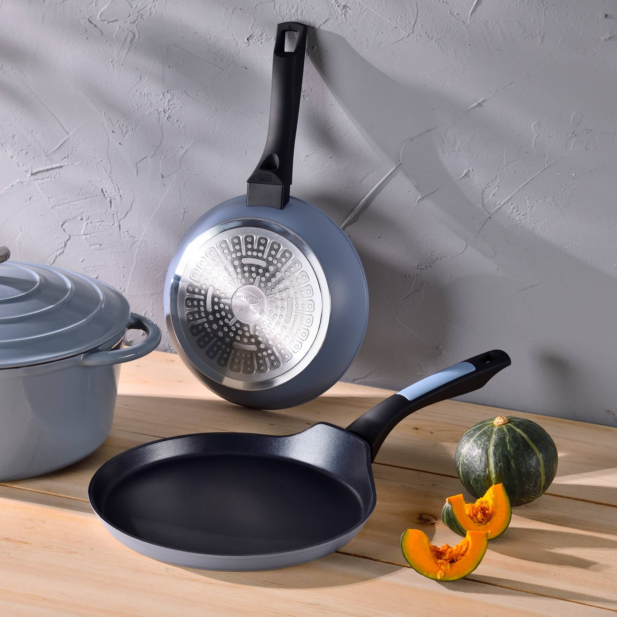 Sartenes y una olla sobre una mesa de madera. Una calabaza entera y dos rebanadas están junto a la sartén. Fondo gris.