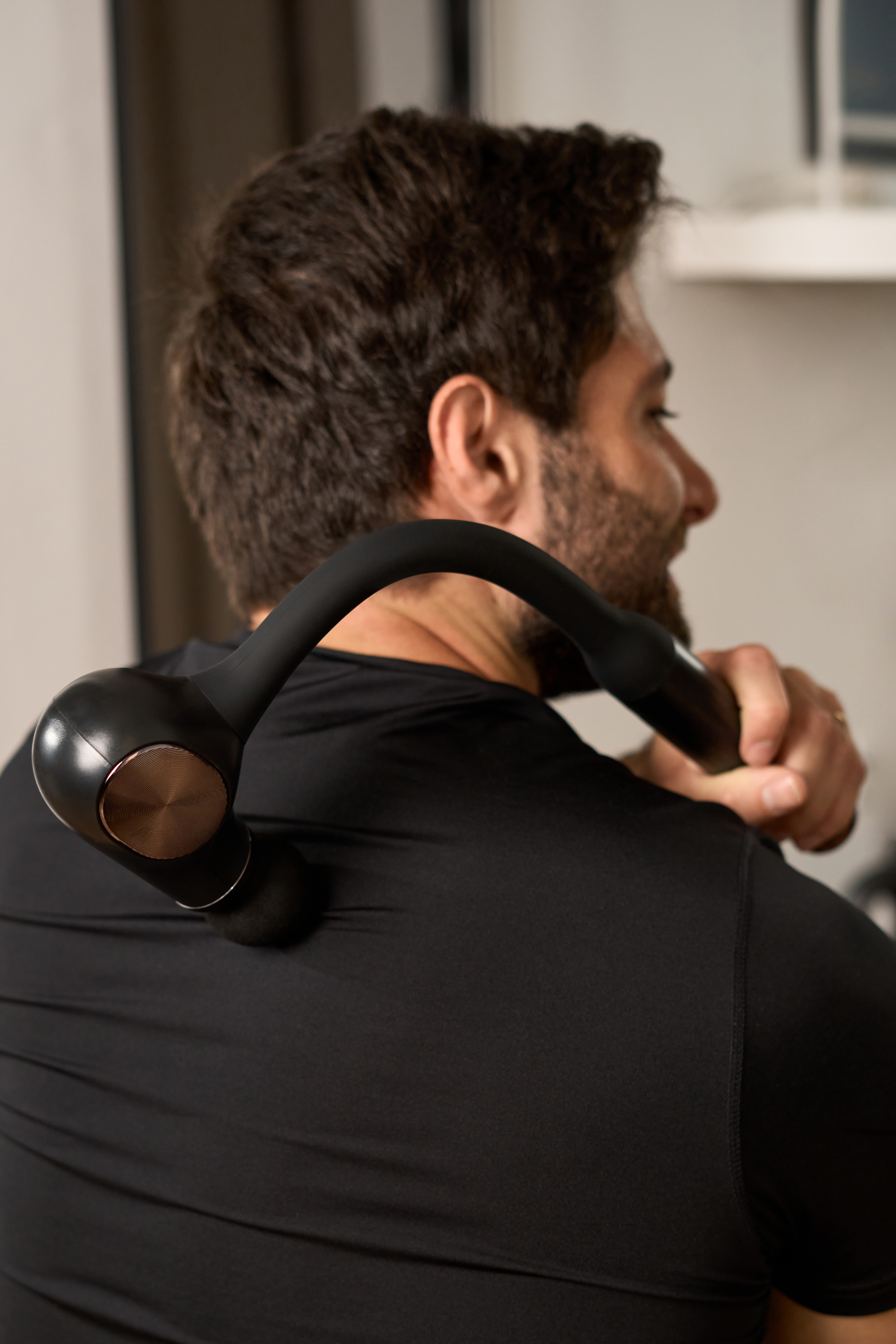 Mężczyzna używa czarnego narzędzia do masażu na plecach. Ma na sobie czarną koszulę. Tło jest rozmyte.