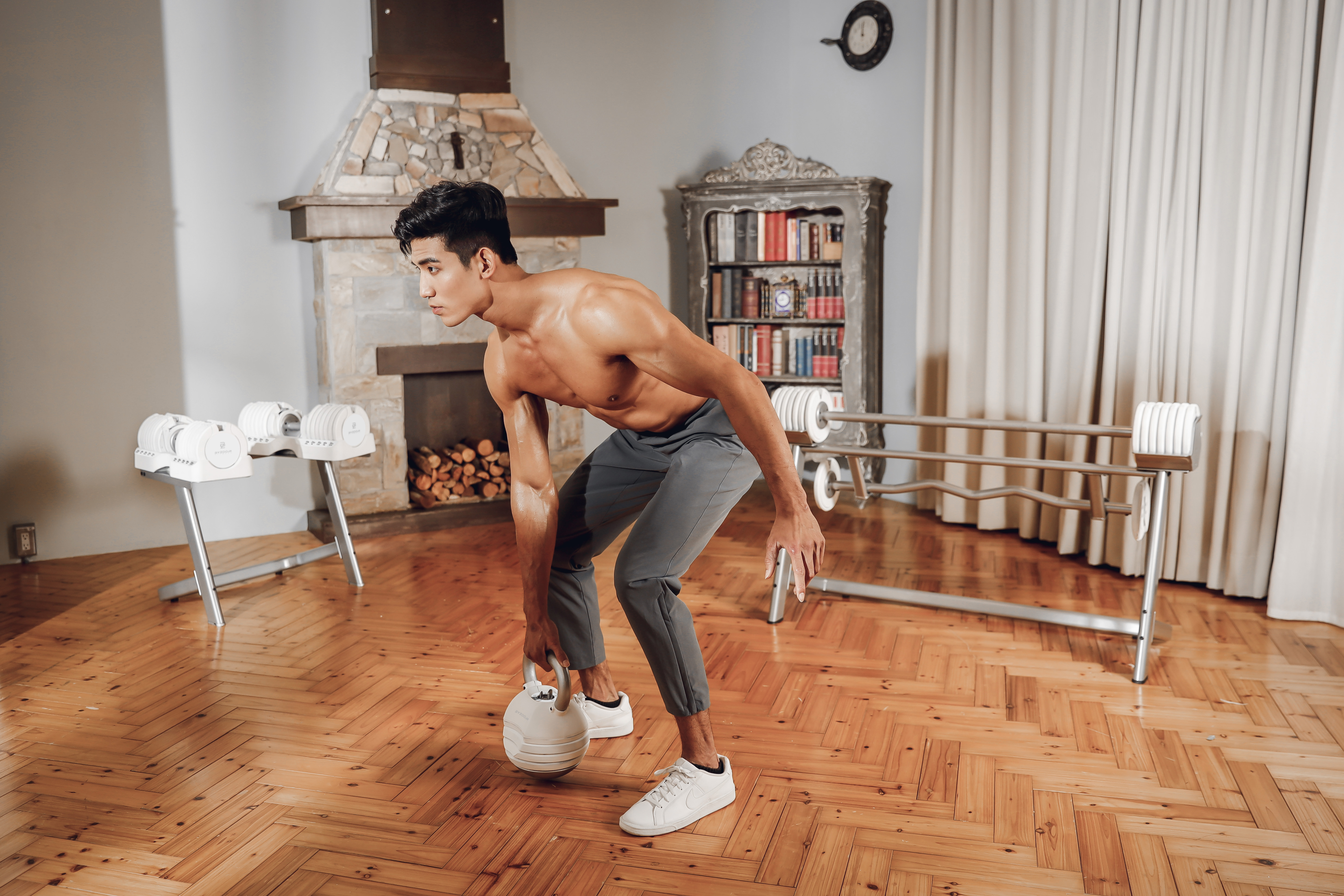 Ein Mann hebt eine Kugelhantel in einem Heim-Fitnessstudio. Kamin, Hanteln und Vorhänge im Hintergrund.