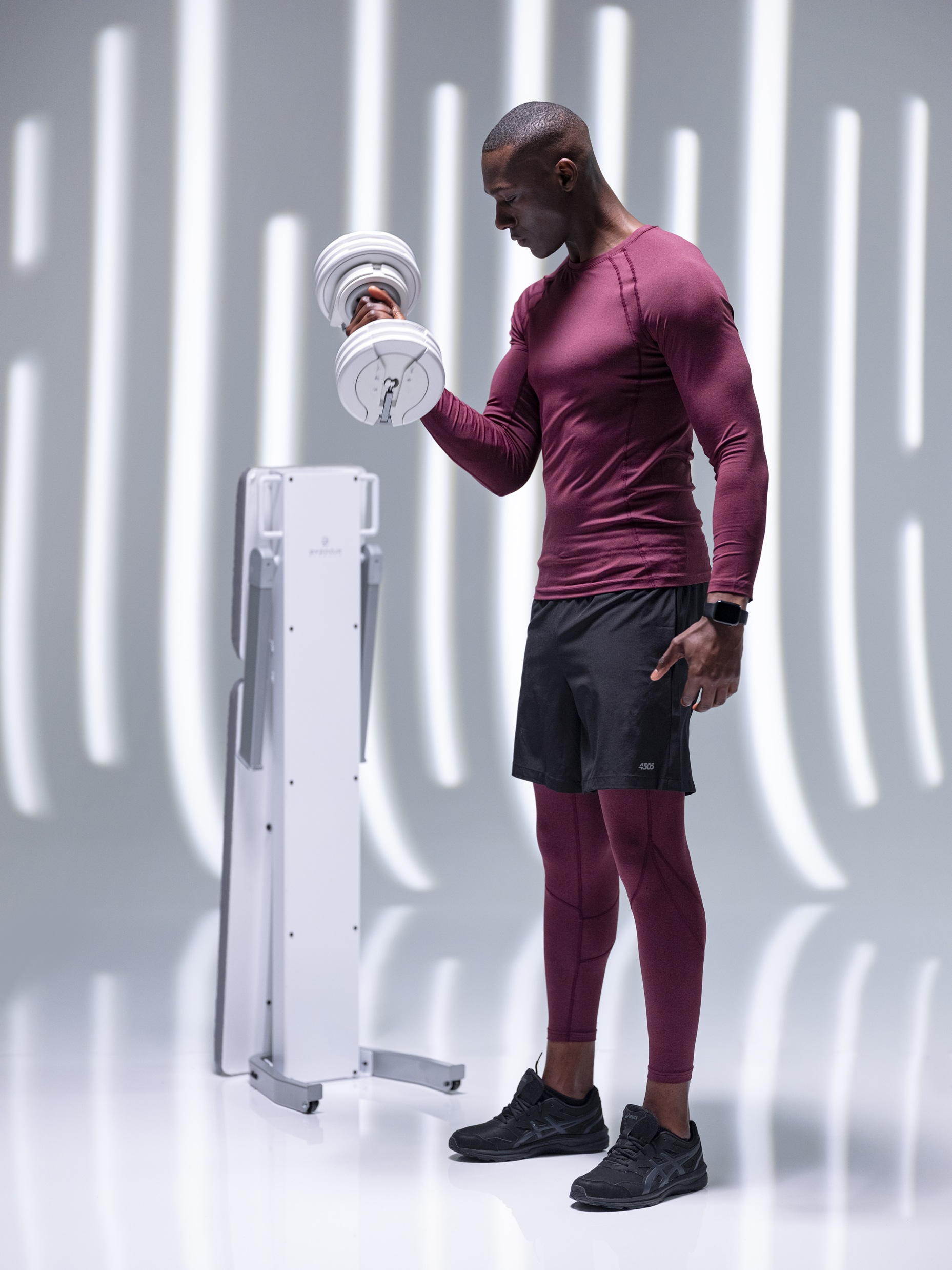 Un homme en chemise et legging bordeaux fait des curls avec un haltère. Une machine blanche est à côté. Fond avec lignes blanches.