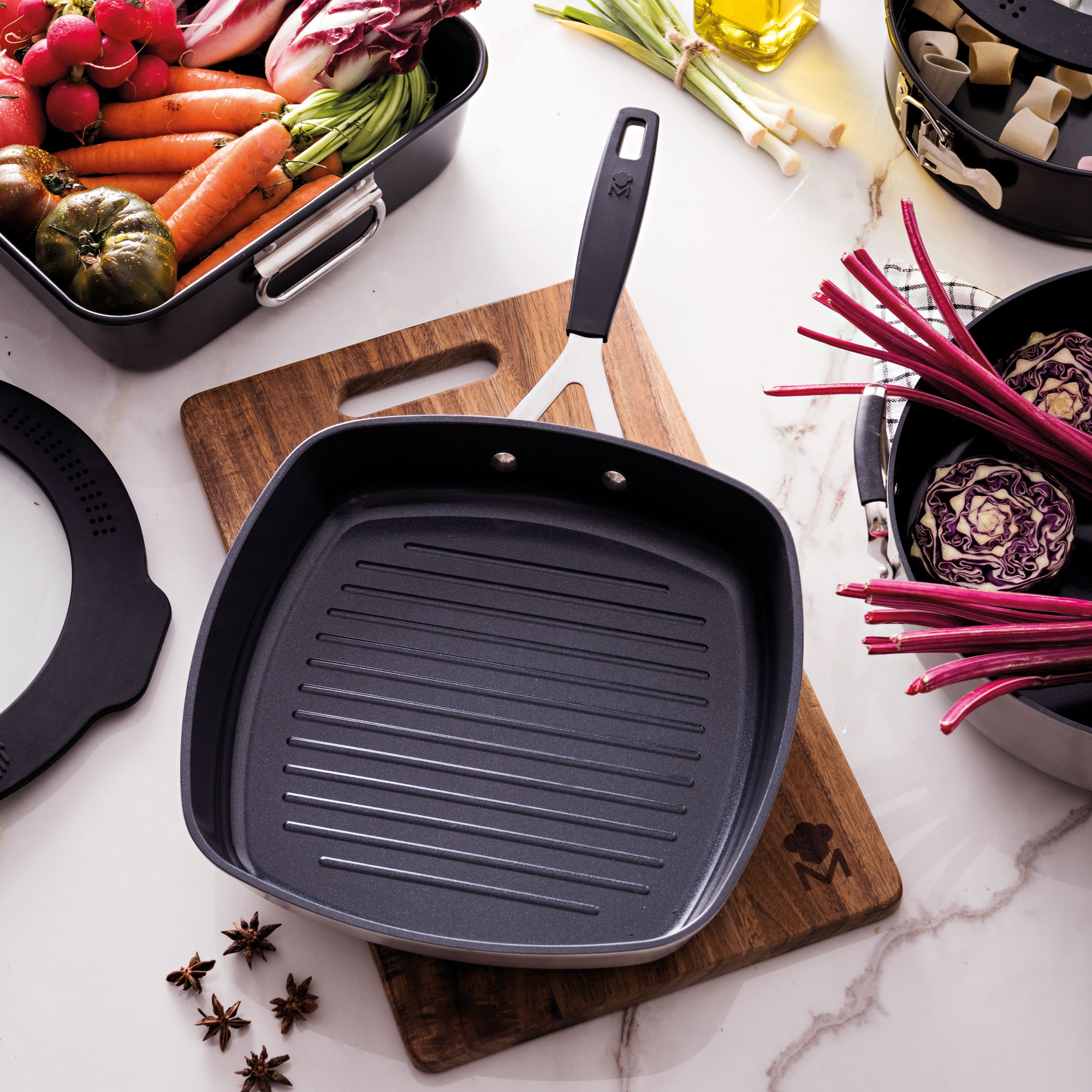 Una sartén para asar a la parrilla sobre una tabla de madera está rodeada de verduras como zanahorias, rábanos y repollo morado, con pasta.