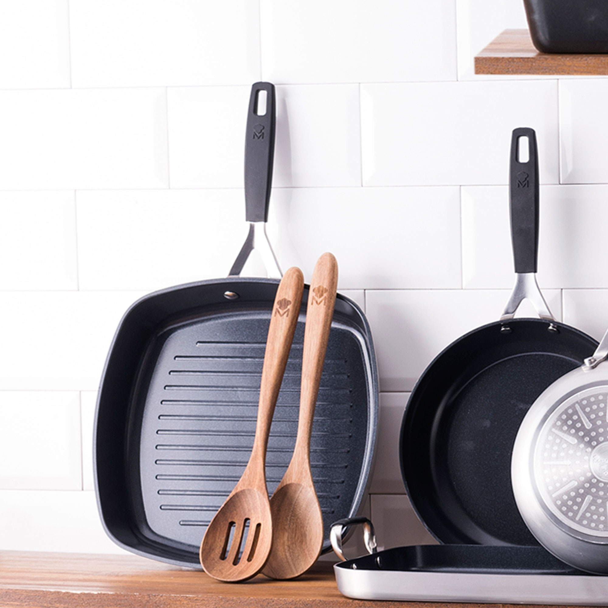 Escena de cocina con sartenes, cucharas de madera y un salpicadero de azulejos blancos. Las sartenes son negras y plateadas. Las cucharas son de madera.