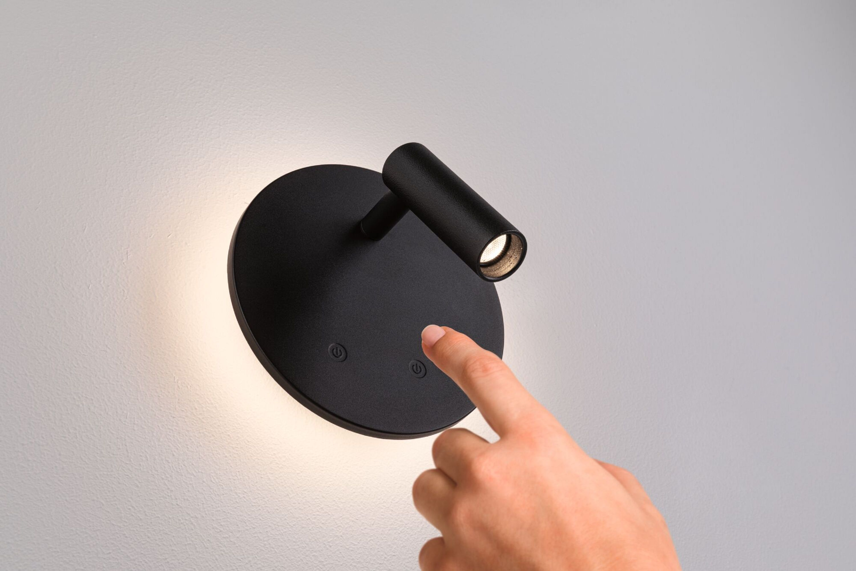 Czarna lampa ścienna z palcem dotykającym przycisku. Lampa jest na białej ścianie.