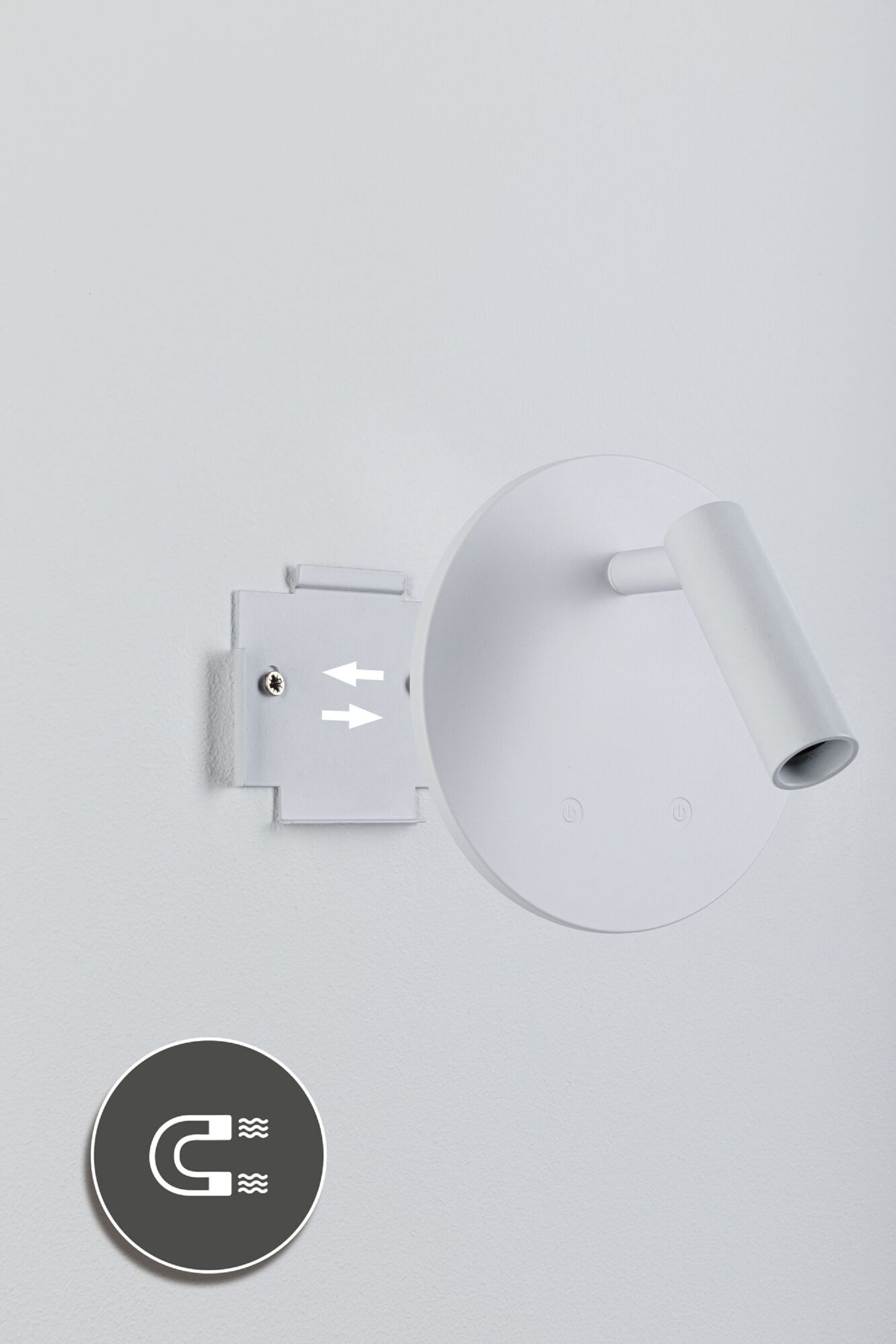 Biała lampa ścienna z okrągłą głowicą i cylindrycznym światłem. Szare koło z symbolem magnesu w rogu.