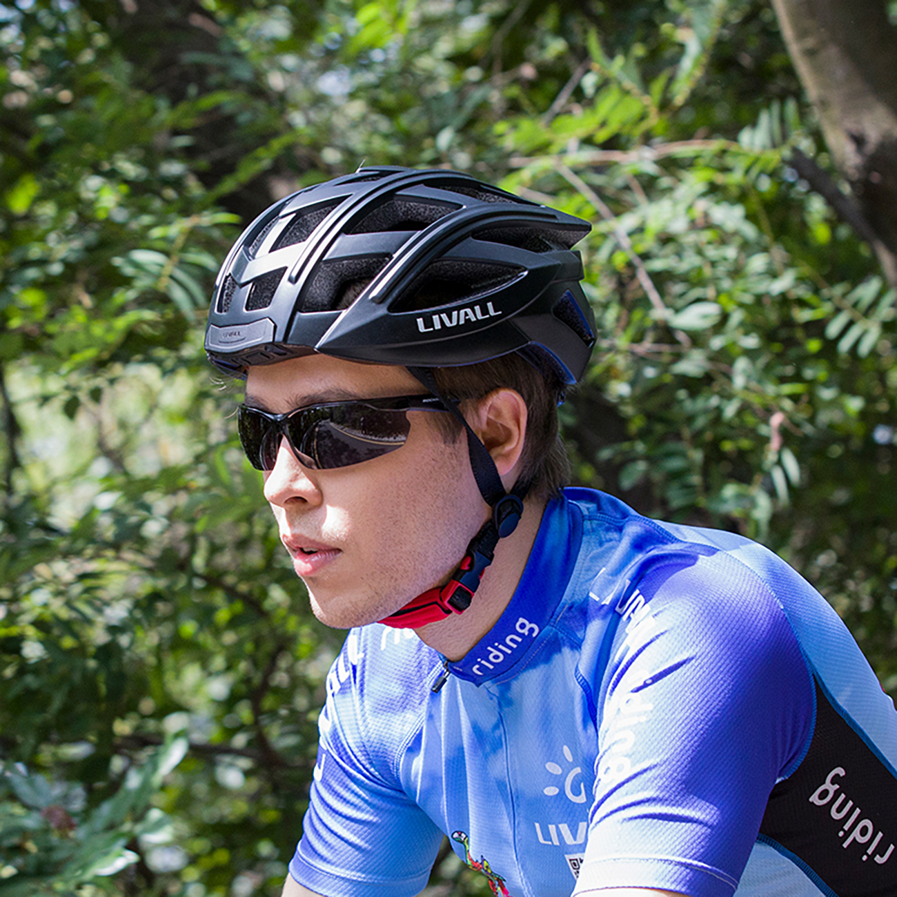 Ein Radfahrer mit schwarzem Helm und Sonnenbrille. Er trägt ein blaues Trikot. Der Hintergrund ist verschwommenes grünes Laub.